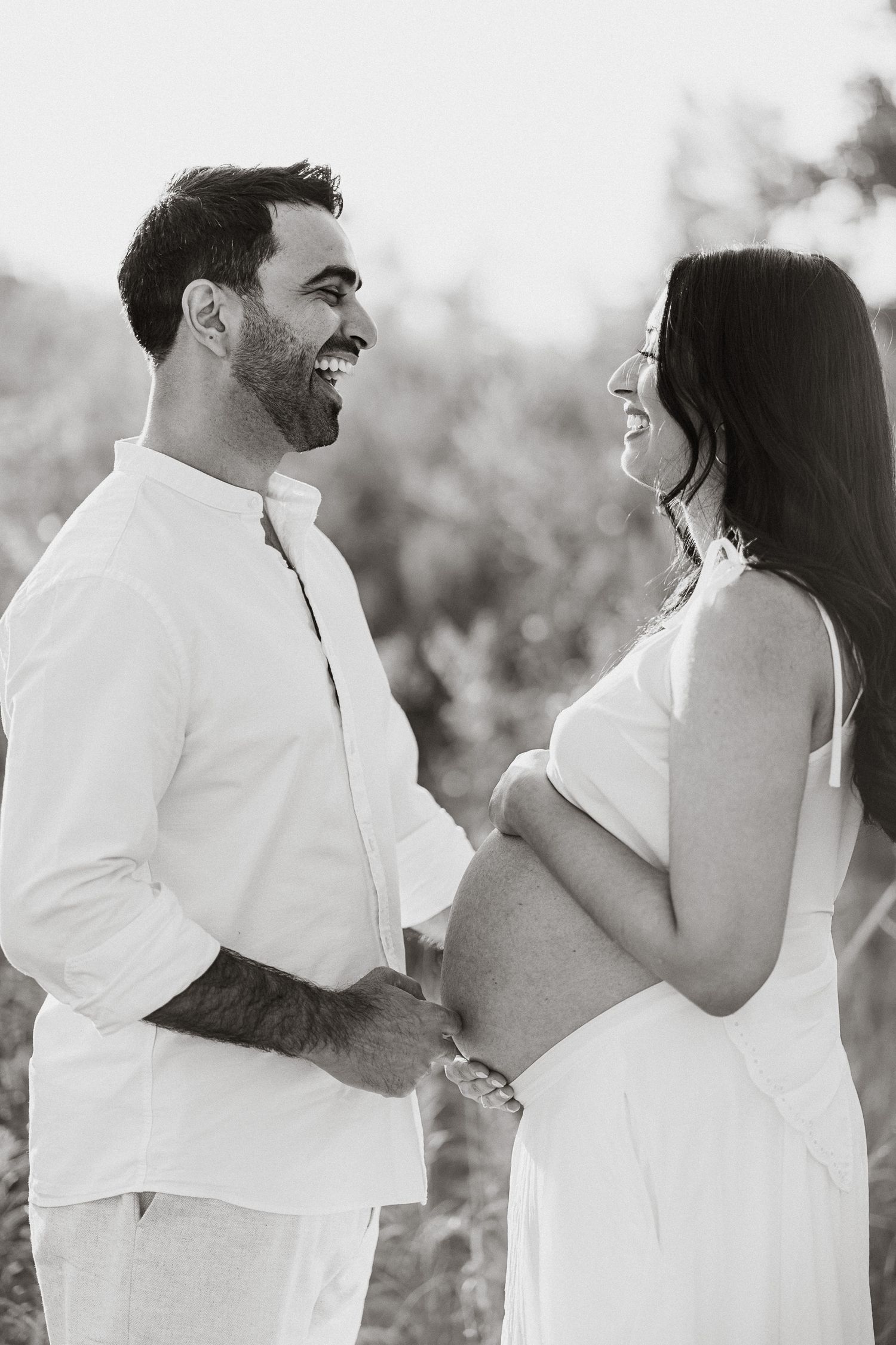 Happy couple in white outfits sharing a joyful moment during a maternity photoshoot outdoors.
