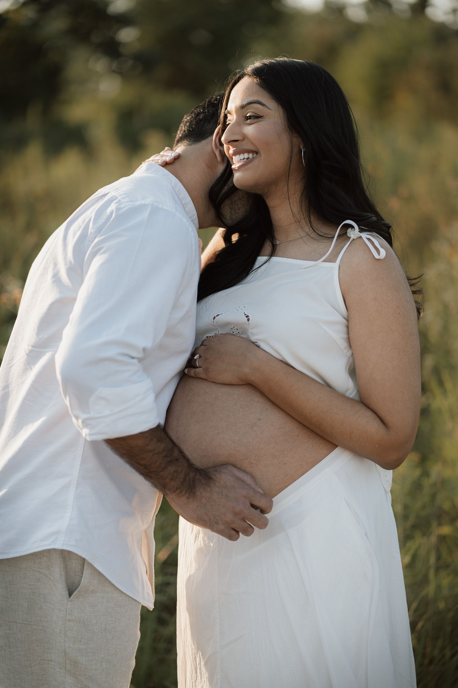 Smiling pregnant woman in white dress embraced by partner outdoors in golden hour light during maternity photoshoot.