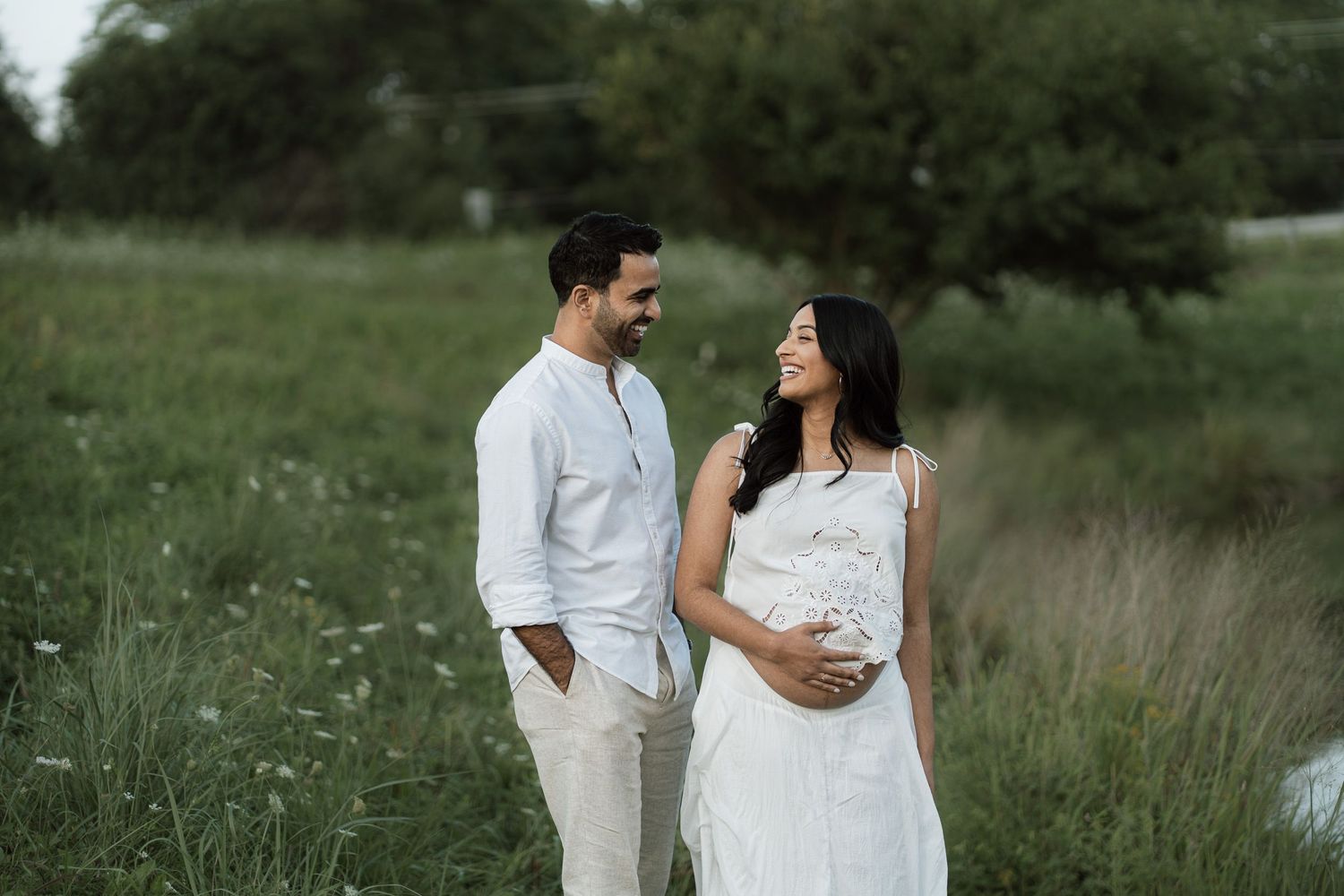 A couple in white attire stands in a green field, smiling at each other while the woman cradles her baby bump.