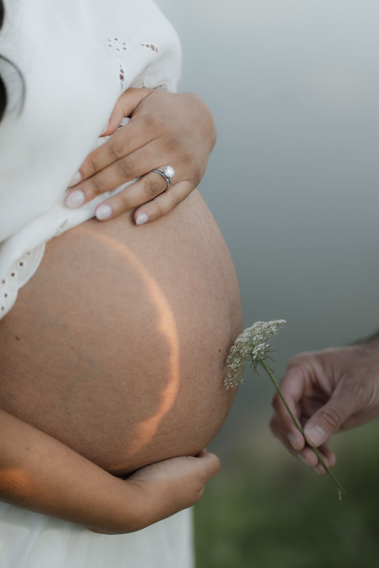Pregnant woman in white dress holding baby bump with diamond ring, as a hand offers a delicate wildflower.