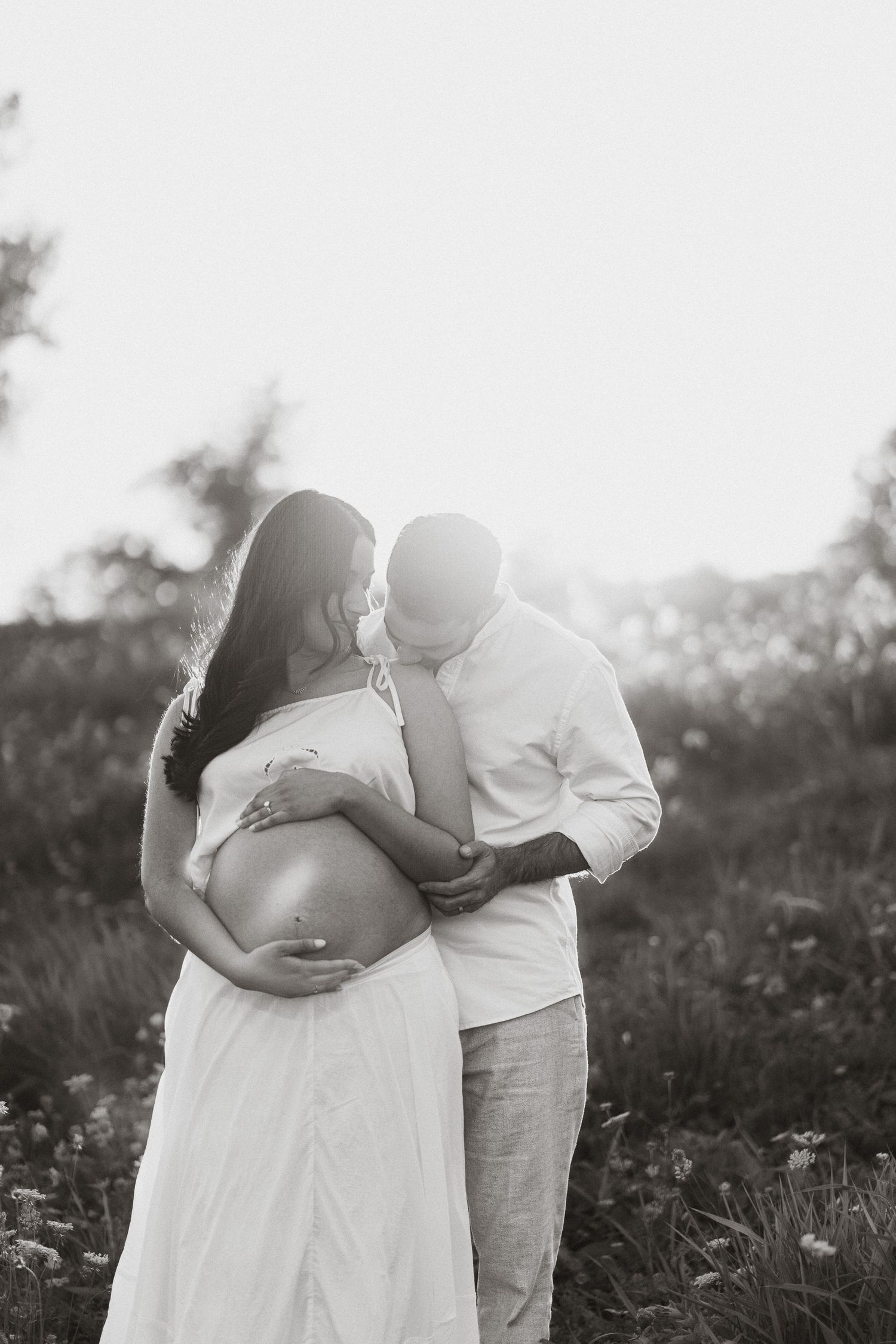 Black and white maternity photo of a couple embracing outdoors, man kissing womans head while she cradles her pregnant belly.