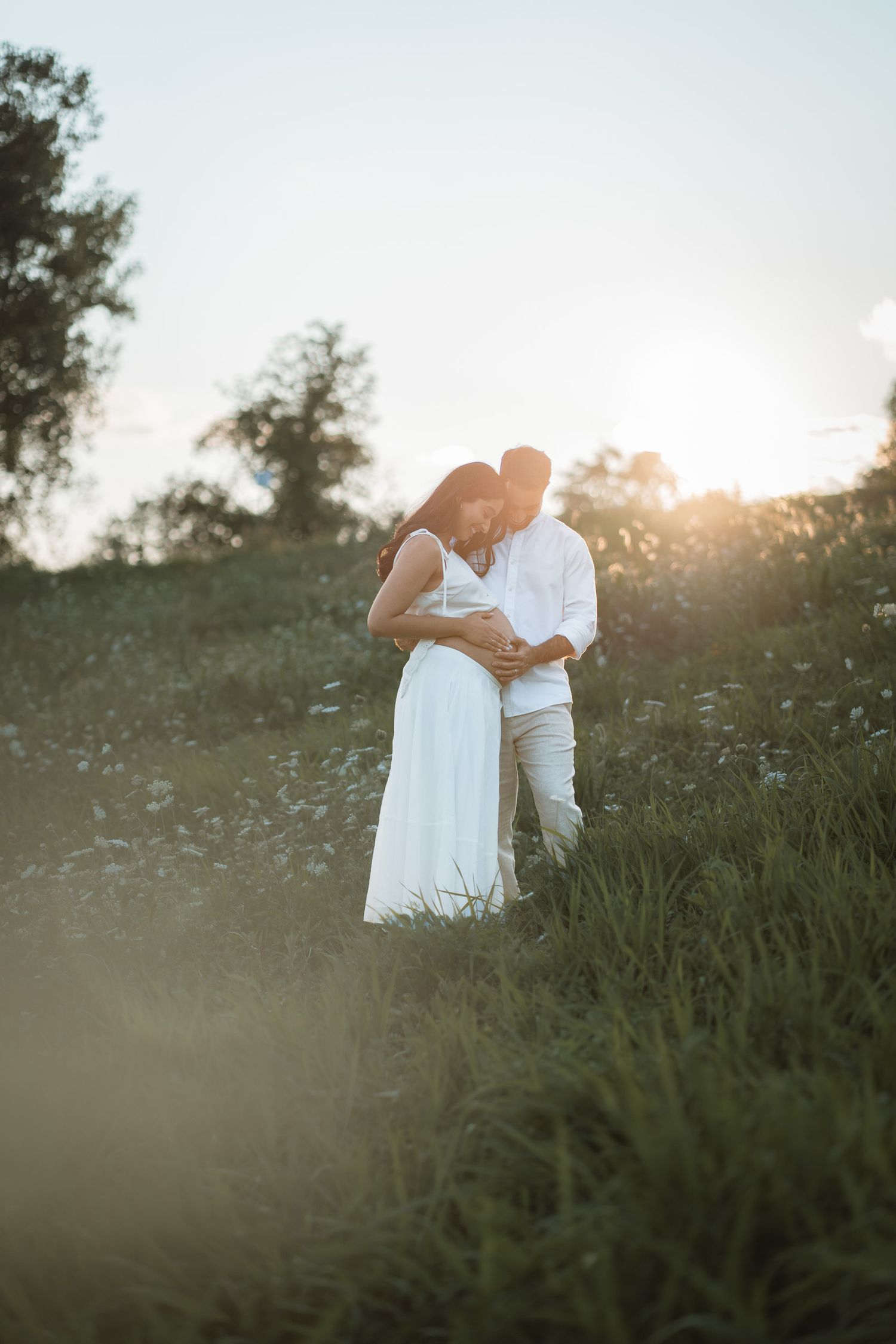 Couple embracing in a sunlit meadow at golden hour, woman in white dress, surrounded by wildflowers and trees.