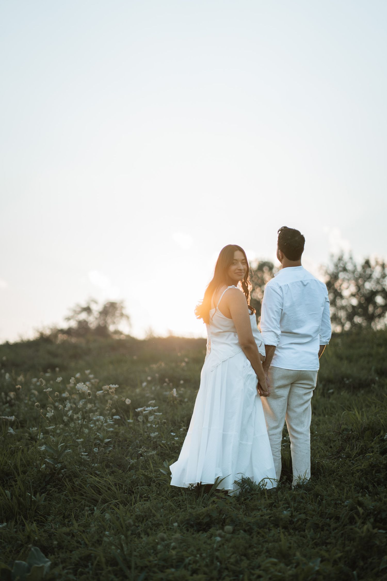 Couple holding hands in a sunlit field at golden hour, woman in white dress looking back over shoulder.