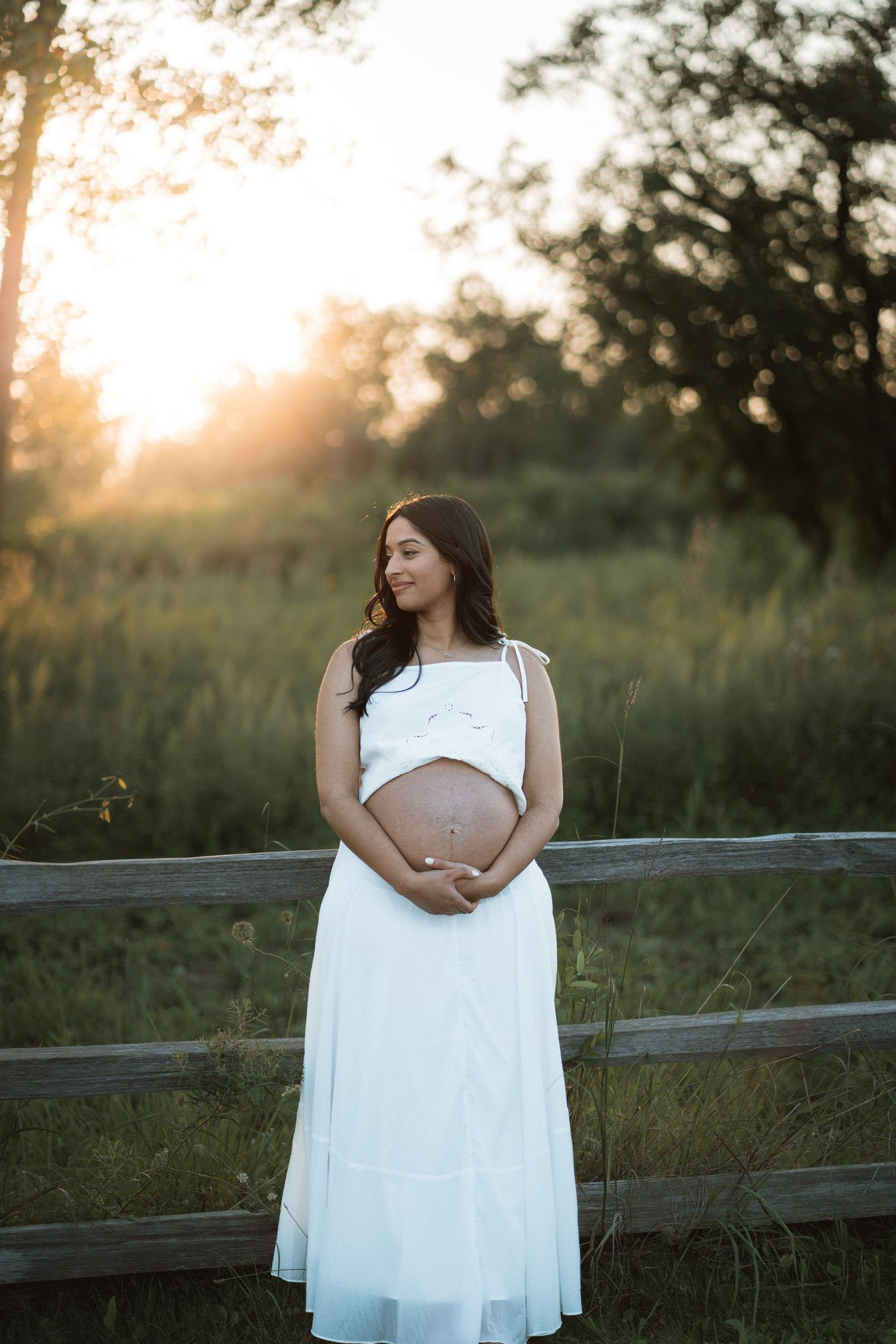 Pregnant woman in white dress cradling baby bump at golden hour near wooden fence in lush green field.
