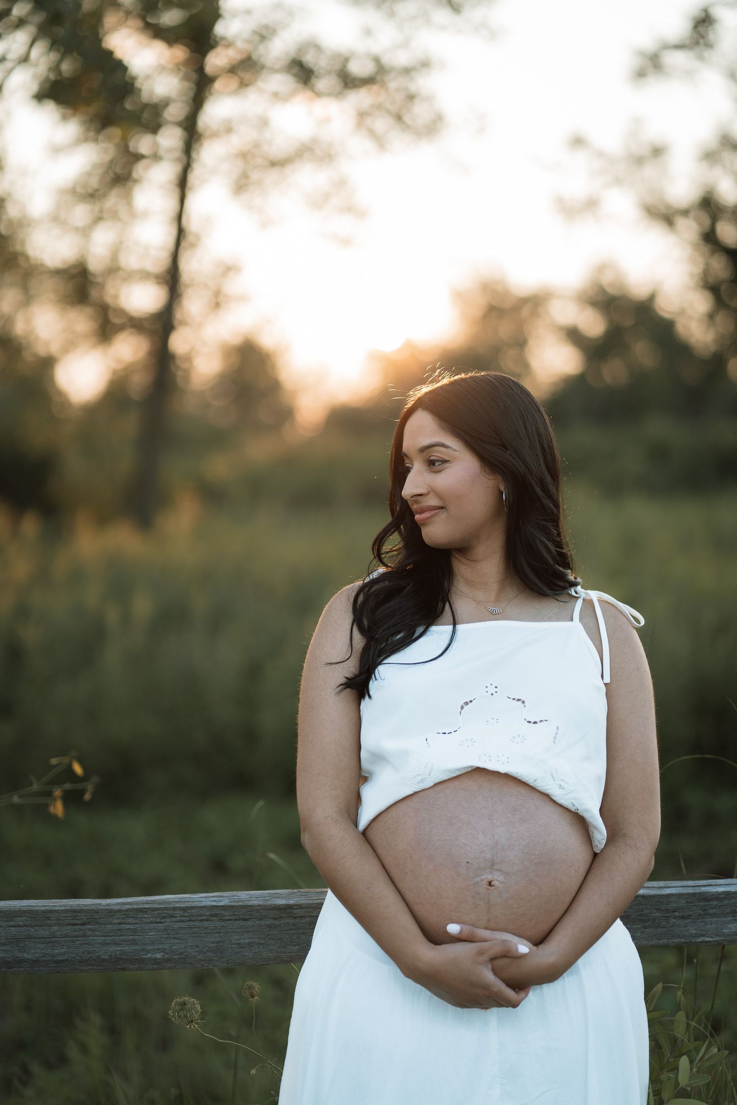 Pregnant woman in white dress cradling baby bump outdoors at golden hour with lush green trees behind her.