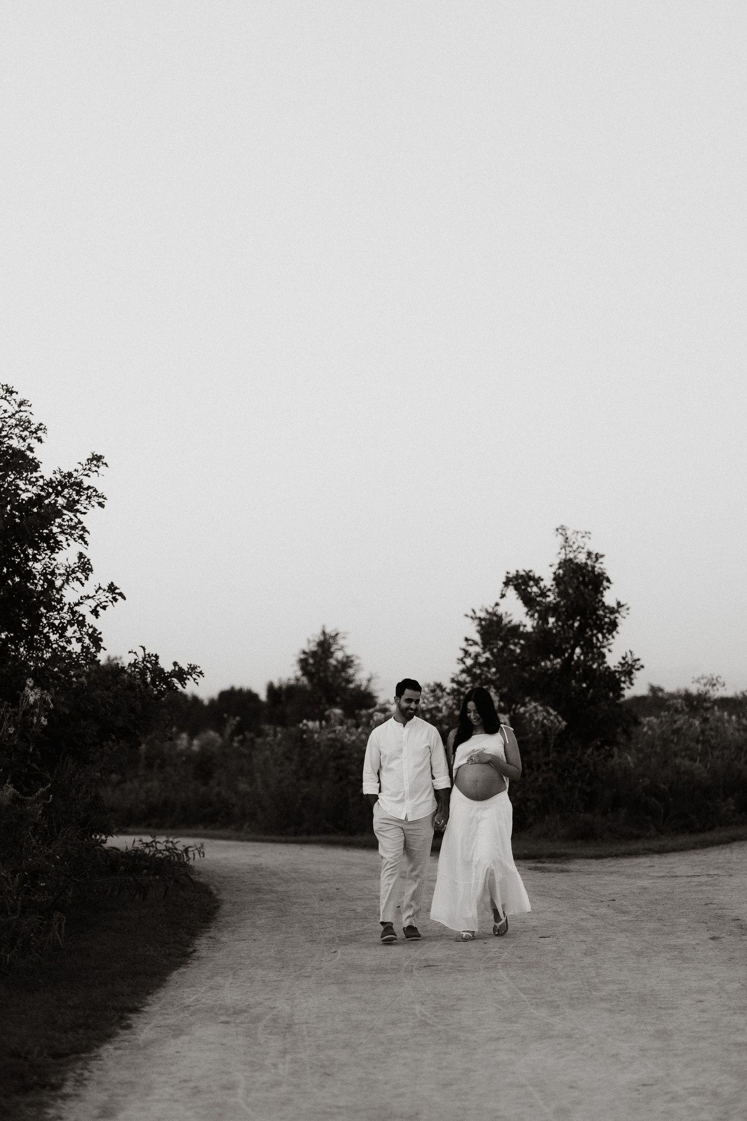 Black and white maternity photo of a couple walking on a path surrounded by trees, woman in white dress showing baby bump.