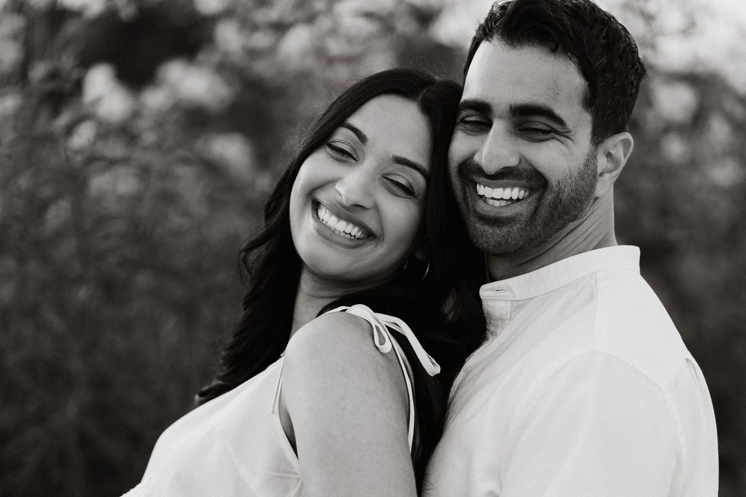 Black and white photo of a smiling couple in white outfits, sharing a joyful moment outdoors.