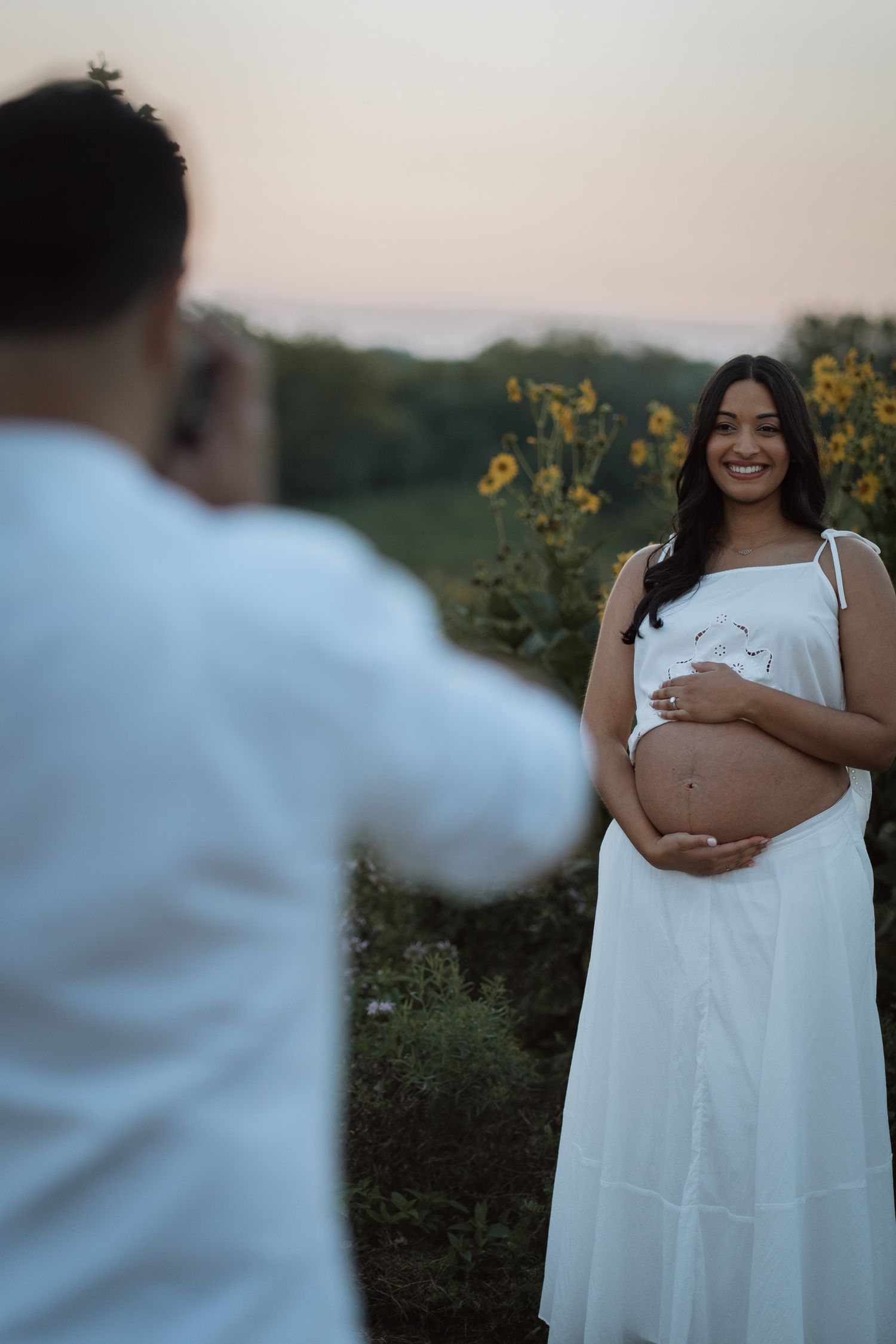 Pregnant woman in white dress smiling outdoors at dusk while partner photographs her bump near sunflowers.