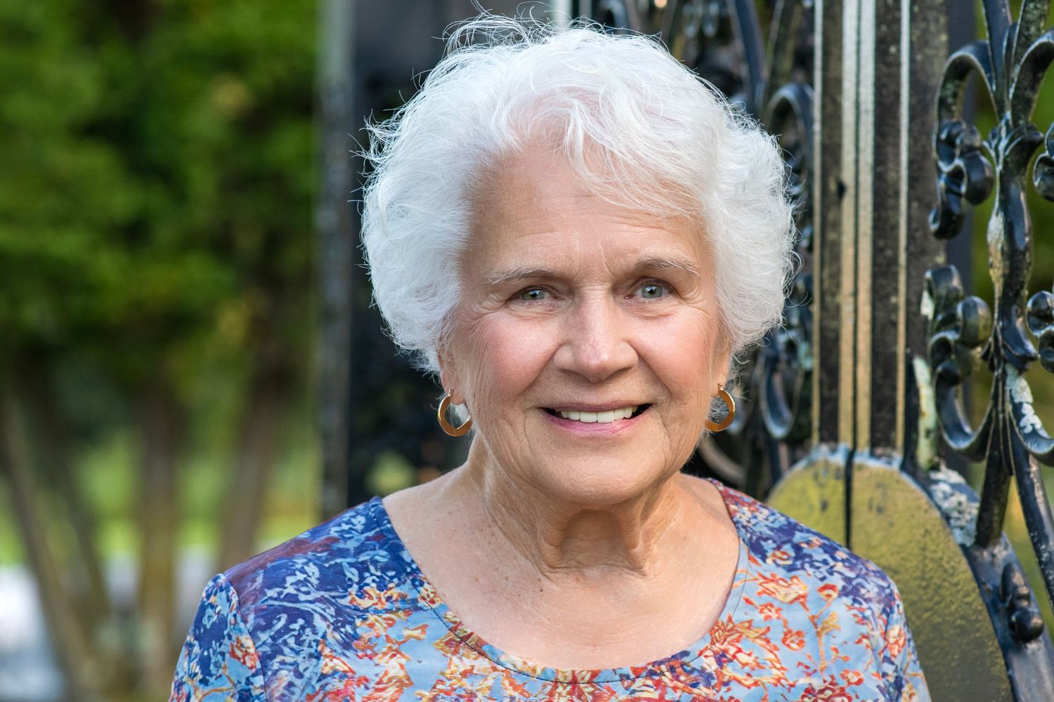 Smiling elderly woman with white hair wearing a floral blouse, standing near an ornate black iron gate outdoors.
