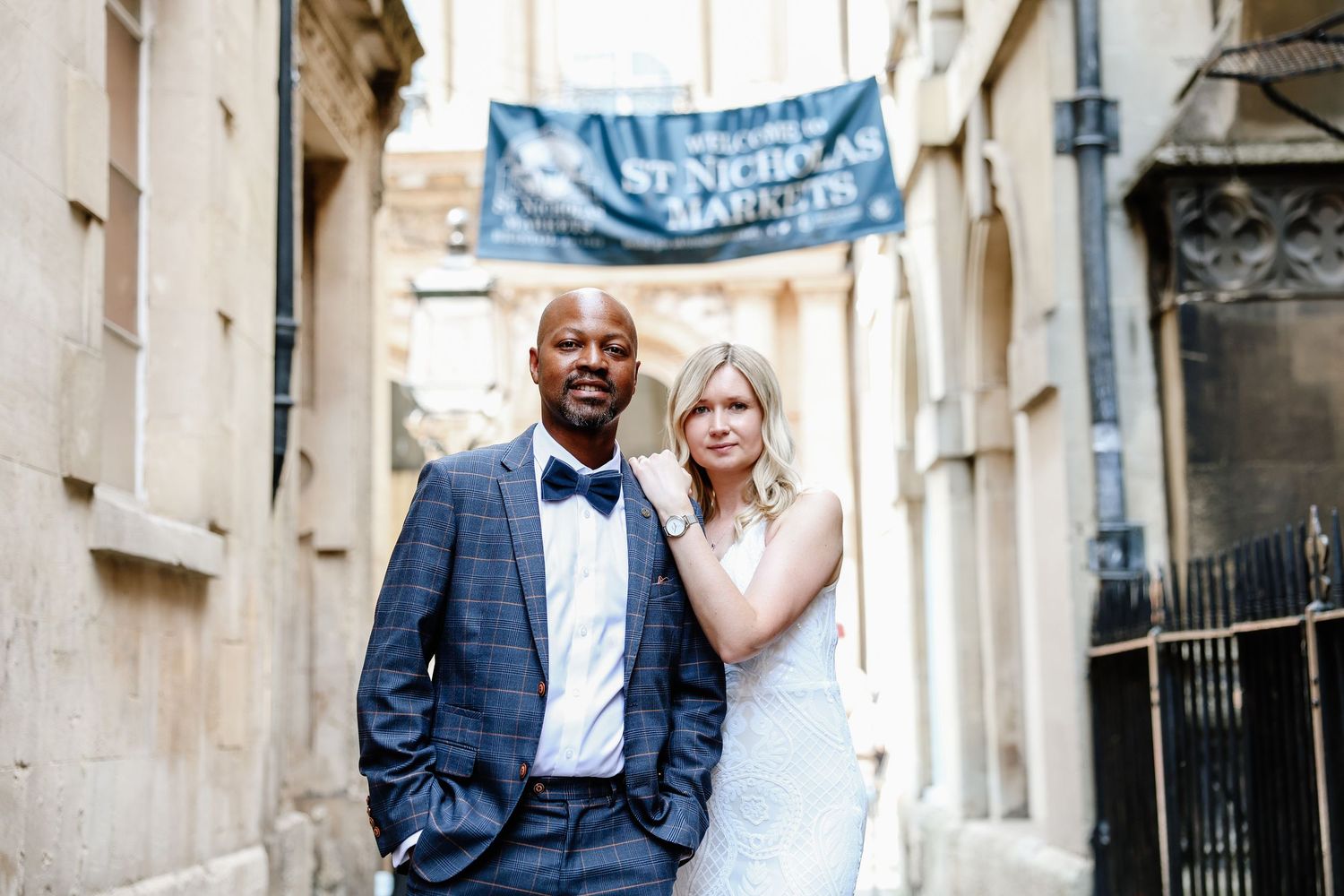 Interracial couple in wedding attire posing in a historic alley near St. Nicholas Markets sign.