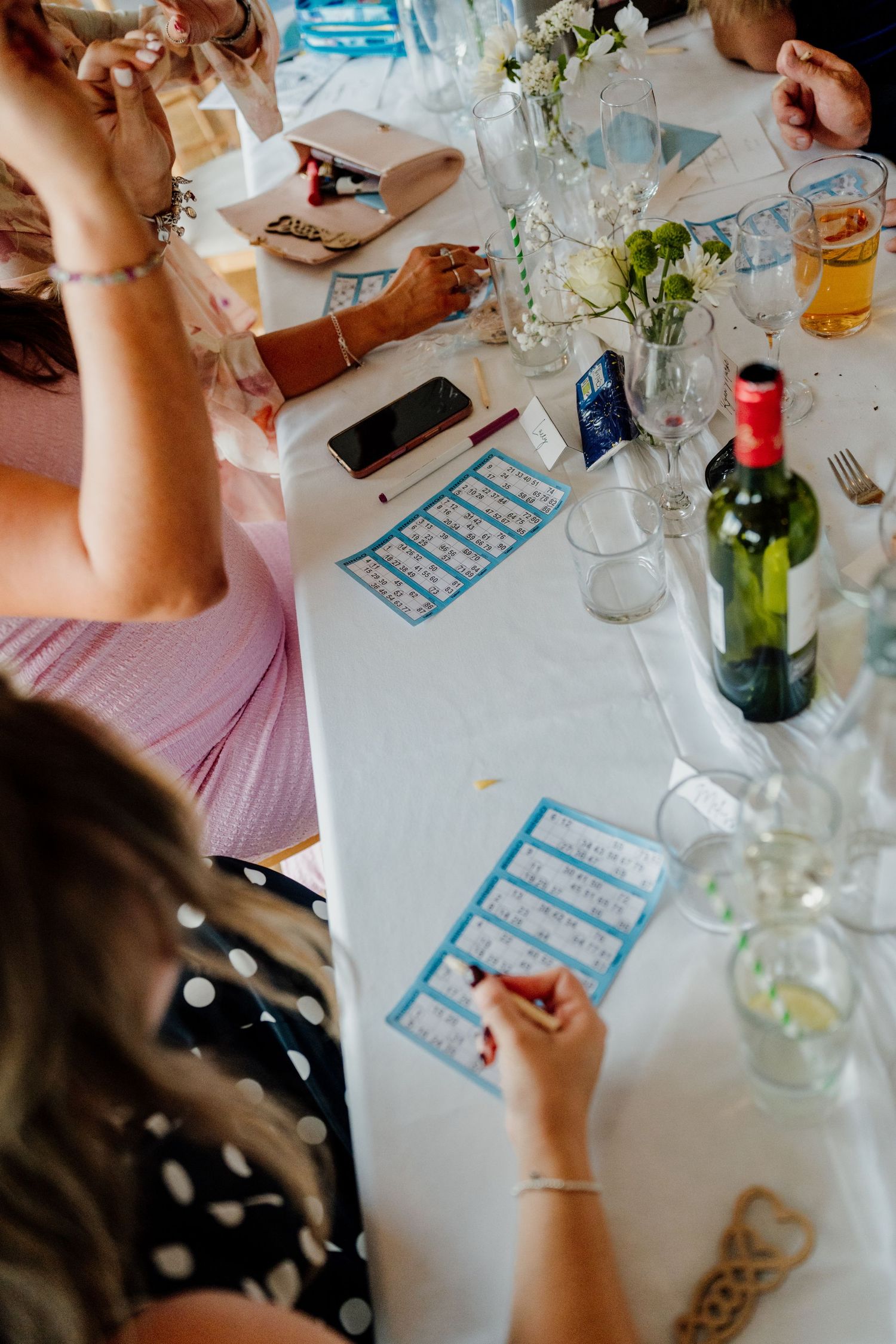 Guests playing bingo at a decorated wedding reception table with wine bottles, glasses, and floral centerpieces.