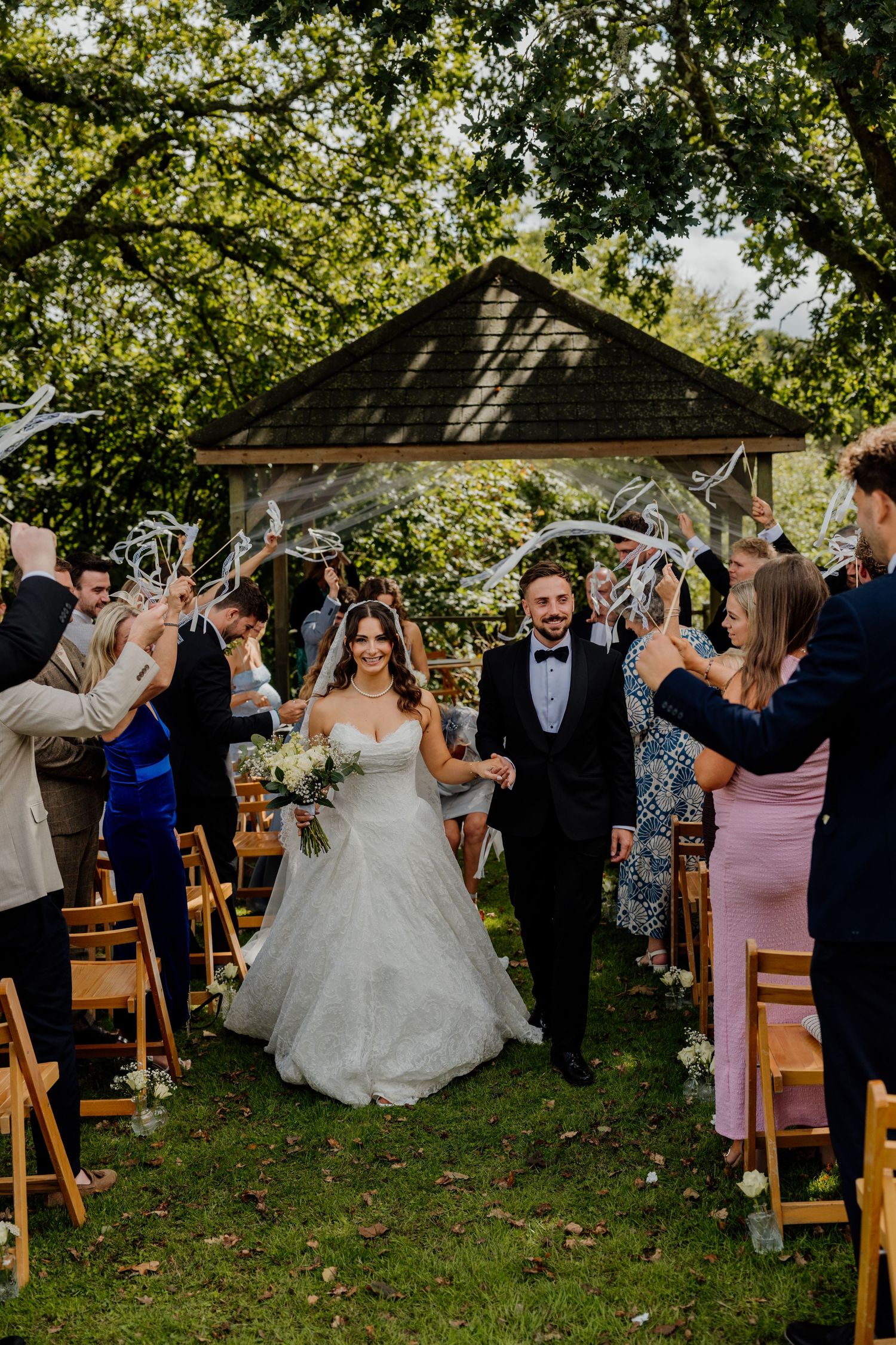 Bride and groom walk down outdoor aisle as guests wave ribbon wands at The Green wedding venue in Liskeard.