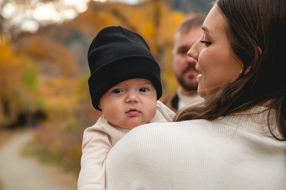 A mother holds her infant wearing a black beanie outdoors in autumn, with a man visible in the background.