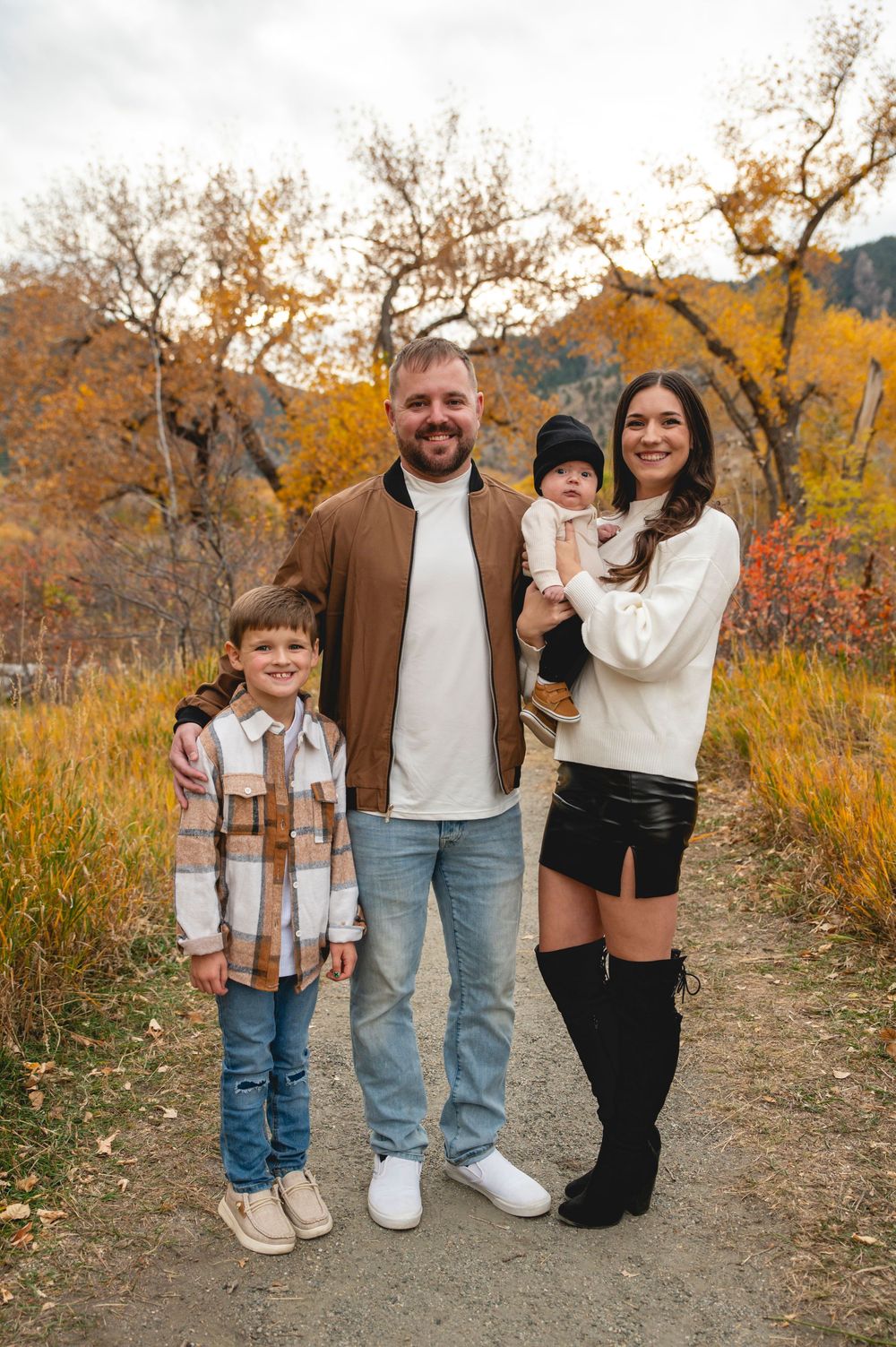 Family of four posing on autumn trail with colorful fall foliage, parents with young boy and baby in seasonal outfits.