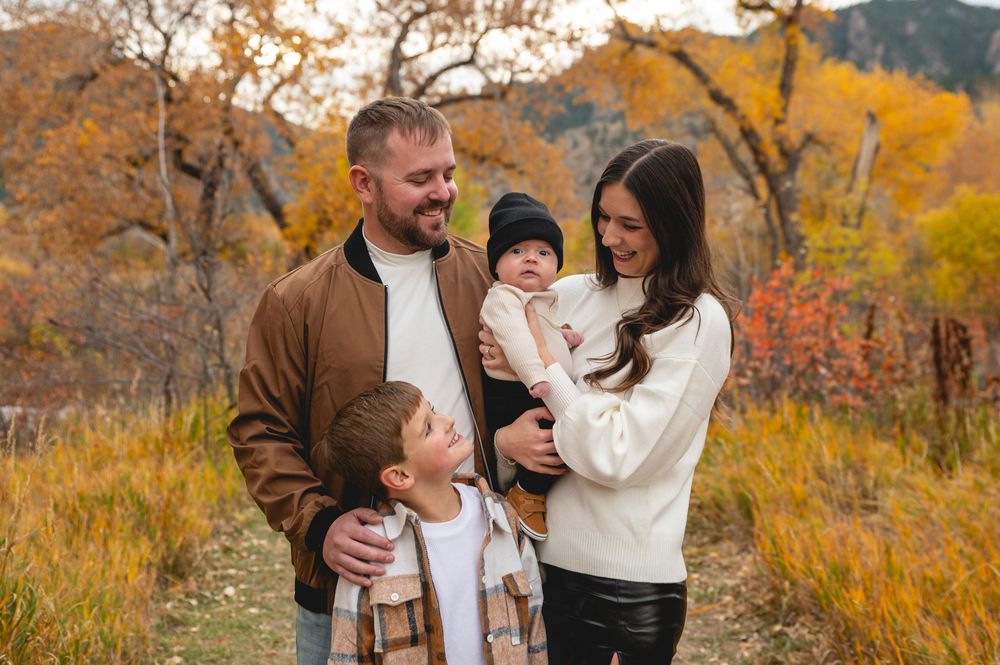 Family of four posing outdoors in autumn foliage, parents smiling with two young children in fall attire.