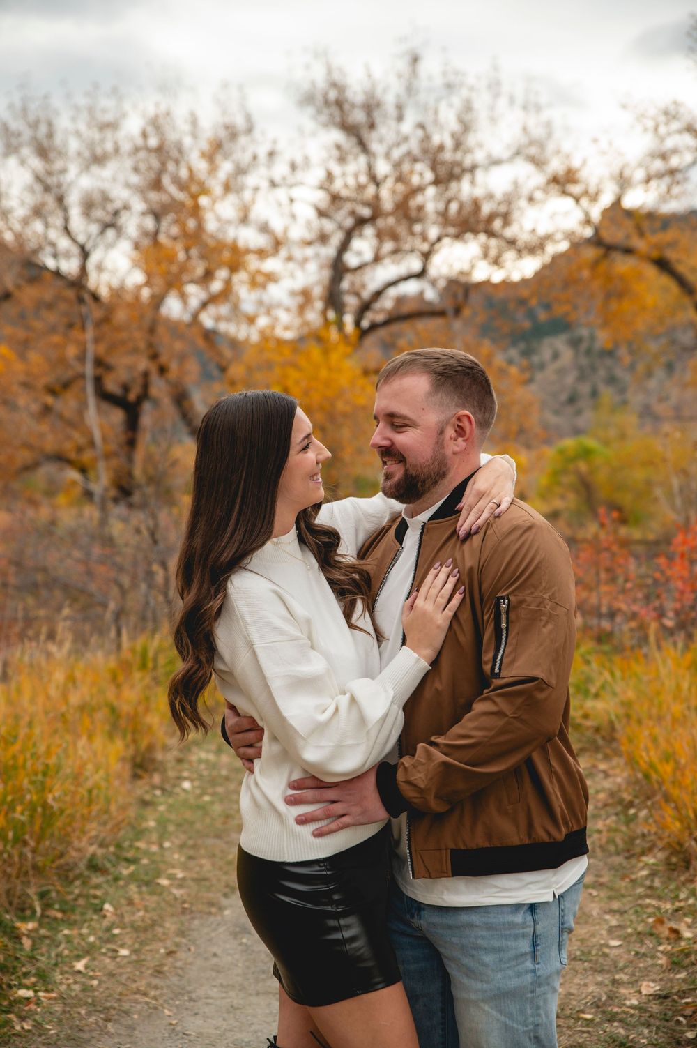Couple embracing on autumn trail, surrounded by golden foliage, woman in white sweater and man in brown jacket.