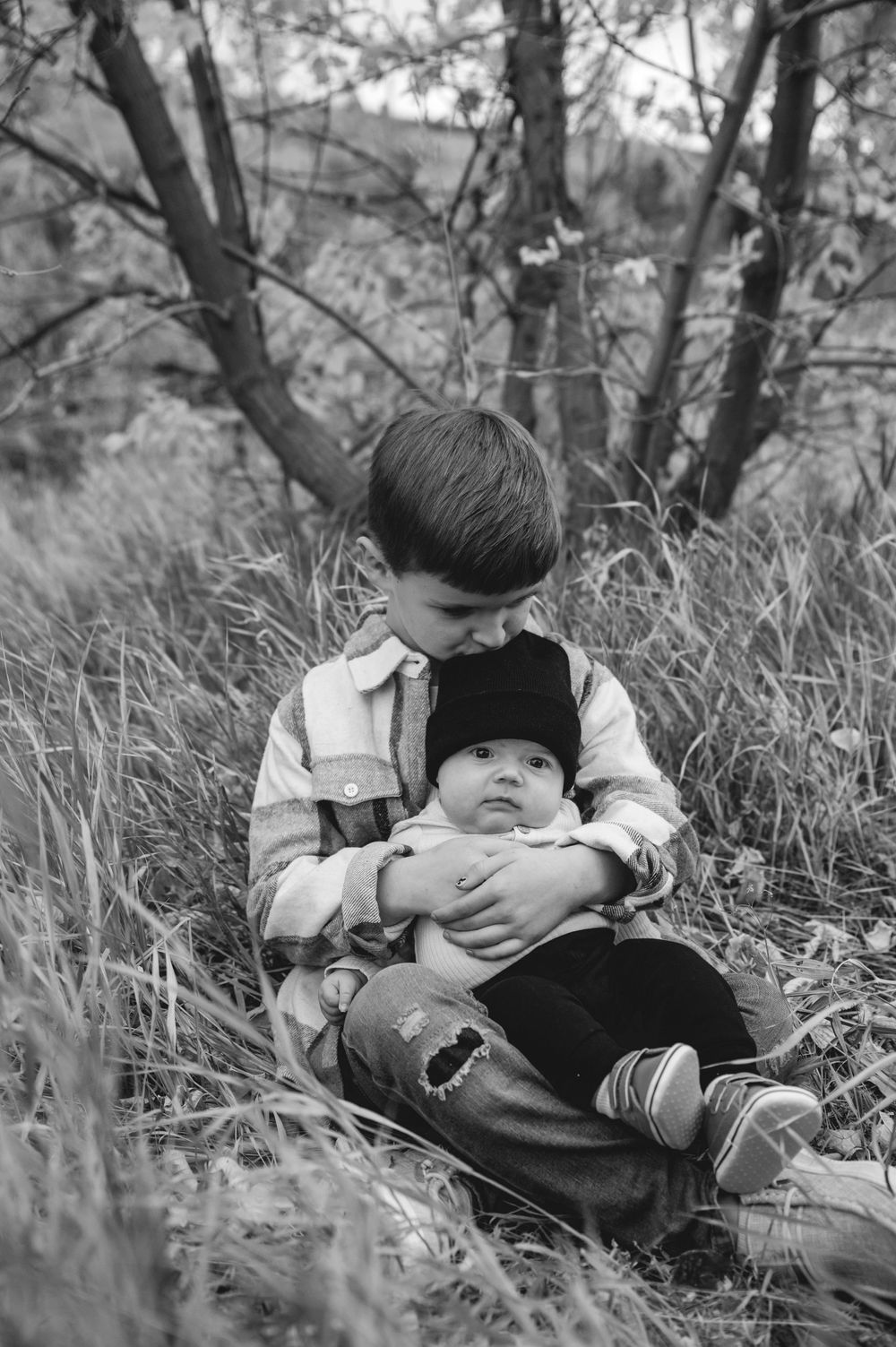 Black and white photo of a young boy sitting in tall grass, gently holding a newborn baby outdoors near trees.