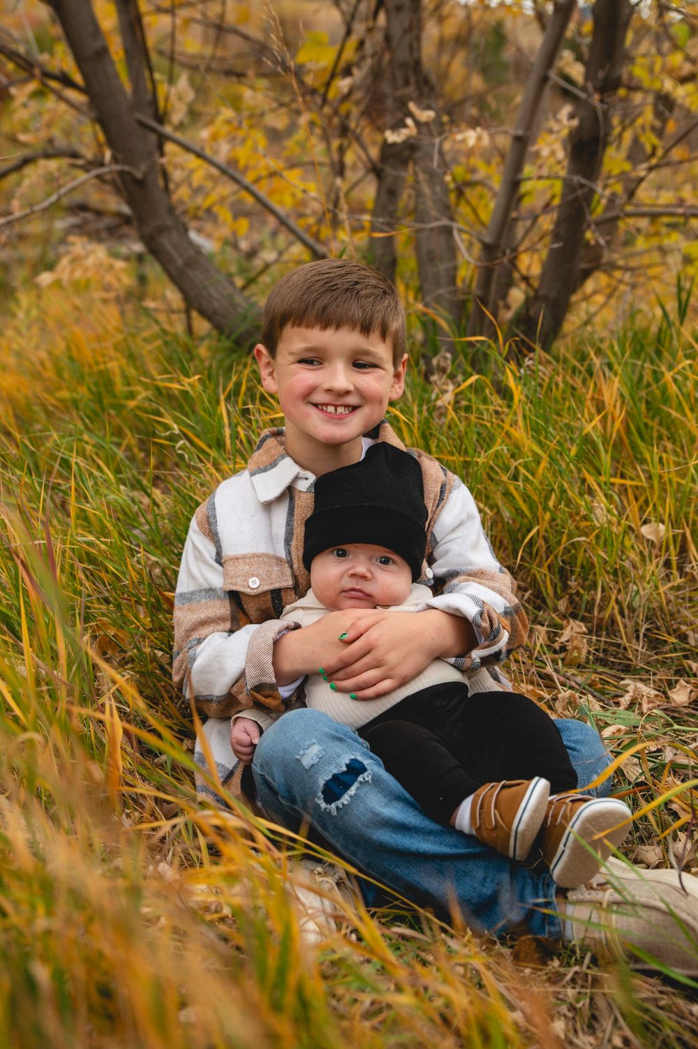 A smiling boy sits in autumn grass holding a newborn baby, both dressed in cozy fall outfits outdoors.