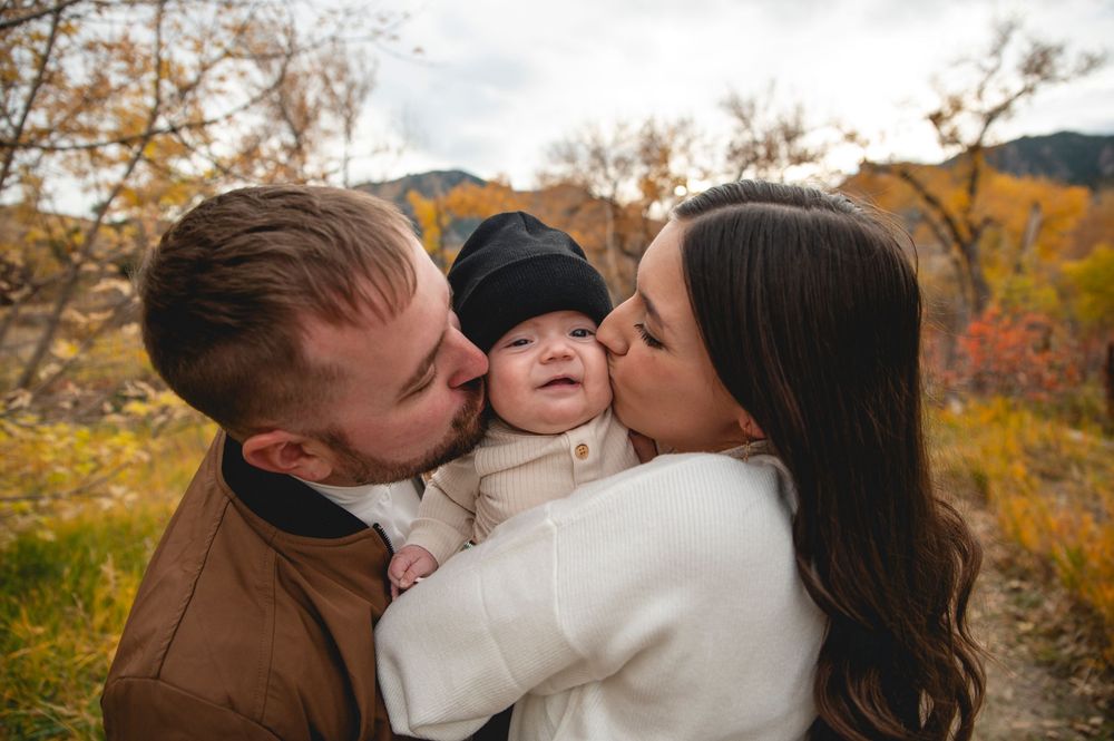 A couple kisses their smiling baby outdoors in autumn, surrounded by colorful fall foliage and mountains.