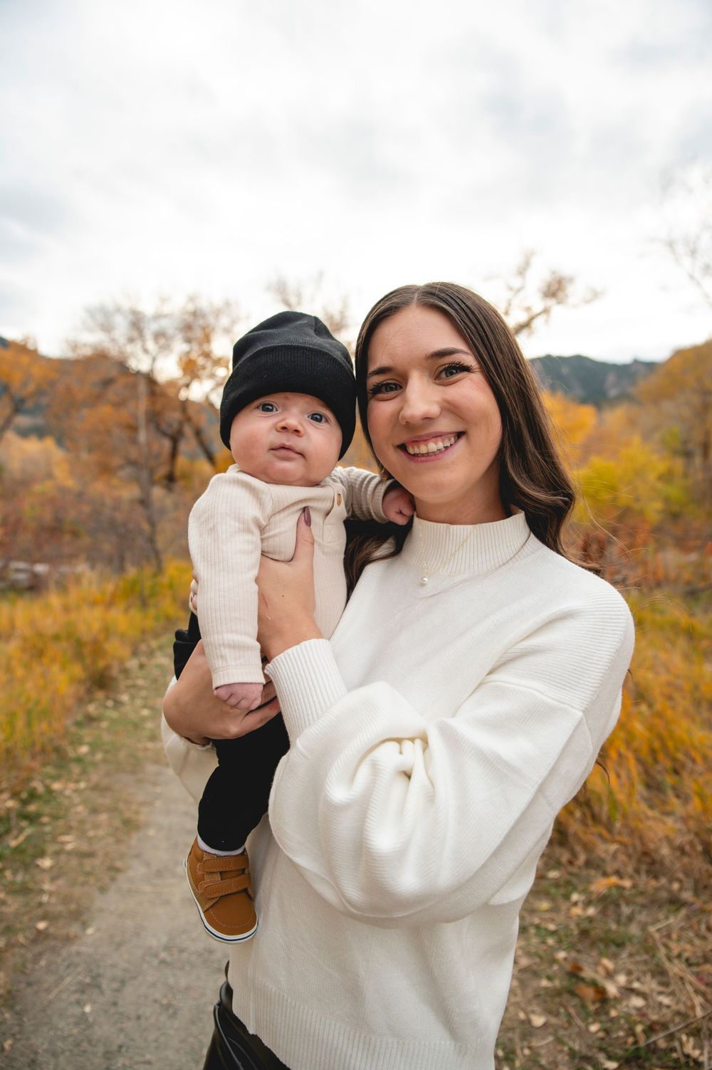Smiling young woman in white sweater holding baby outdoors in autumn landscape with golden foliage.