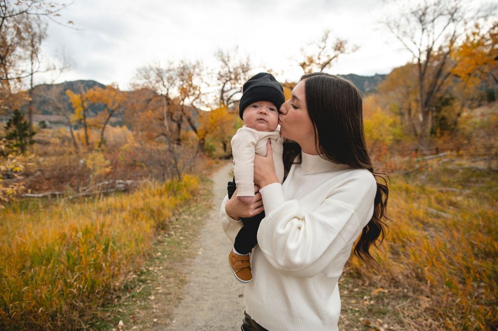 A mother in a white sweater kisses her baby on a scenic autumn trail surrounded by golden foliage.