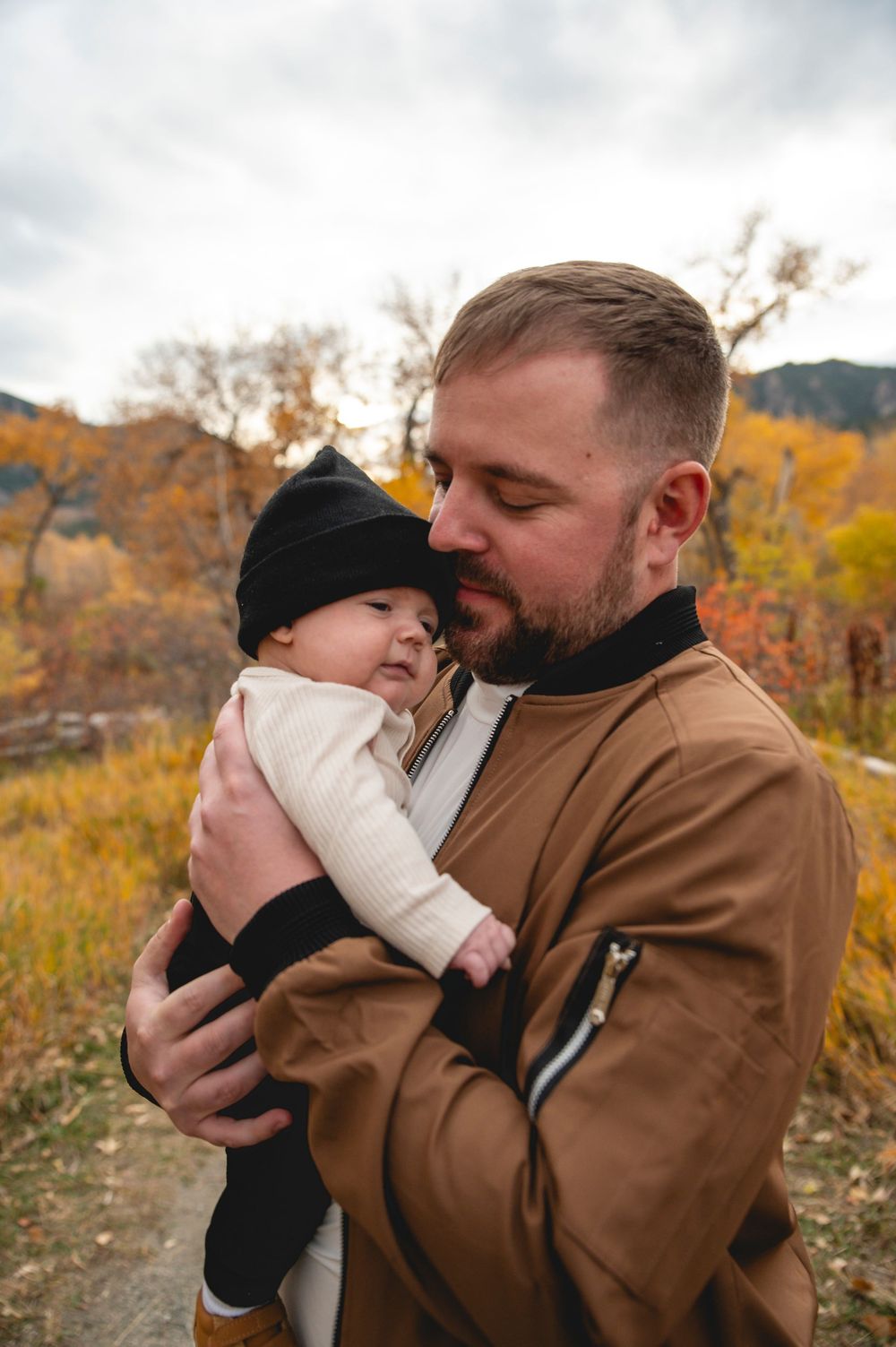 Father in brown jacket tenderly holds infant in black beanie amid autumn foliage outdoors.