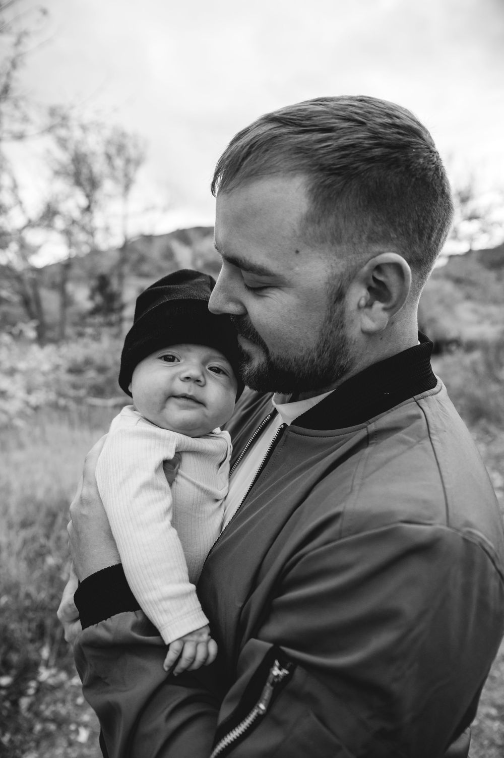 Black and white photo of a bearded man tenderly holding a baby in a hat outdoors.