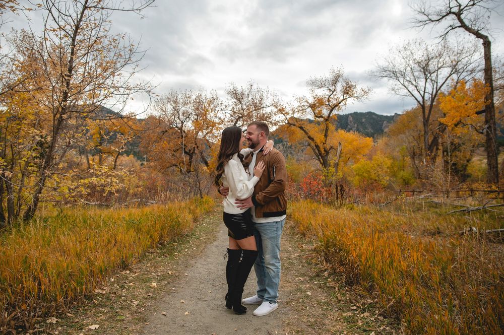 Couple kissing on a scenic autumn trail surrounded by golden foliage and mountain backdrop.