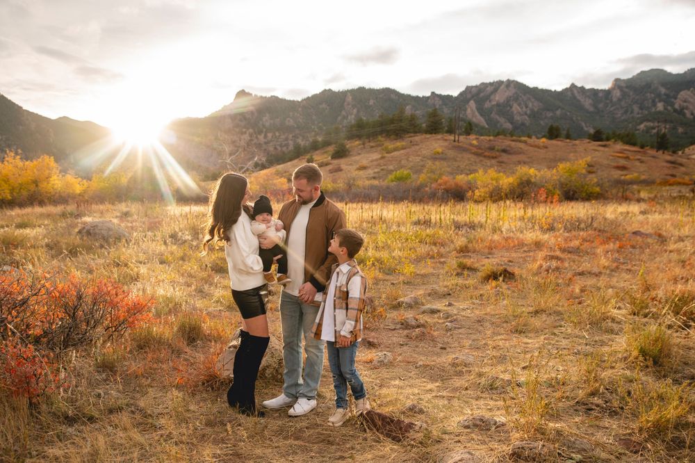 Family of four poses in golden autumn field with mountain backdrop and sunburst at sunset.