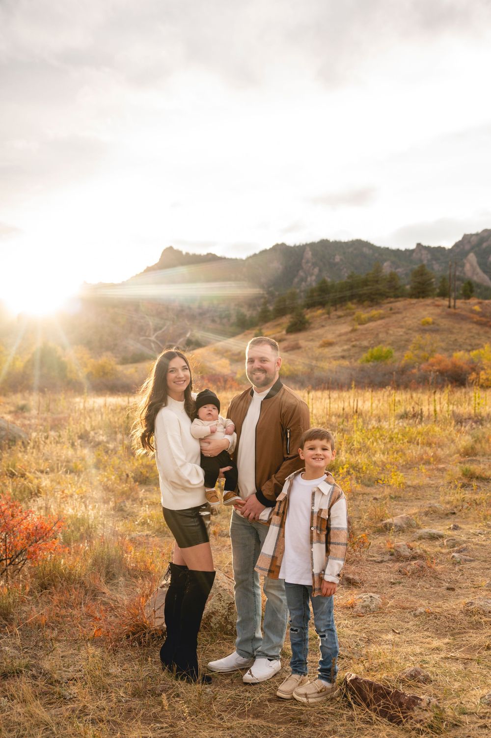 Family of four posing in golden autumn field with mountain backdrop and sunburst, wearing casual fall outfits.