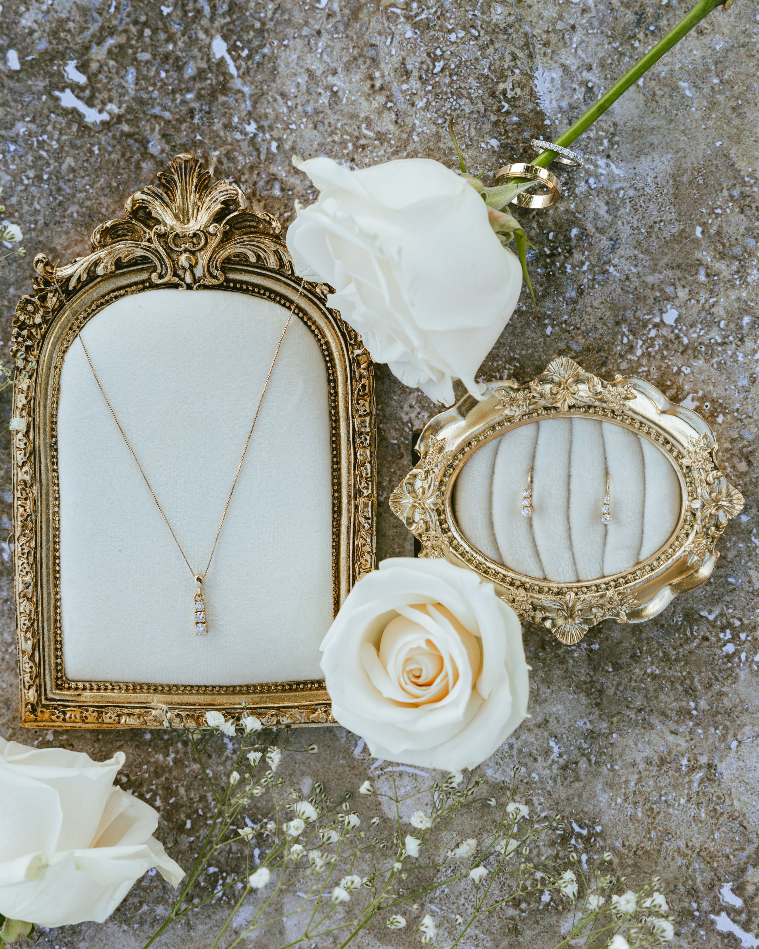 Gold jewelry display with diamond necklace and bracelets in ornate frames surrounded by white roses on stone surface.