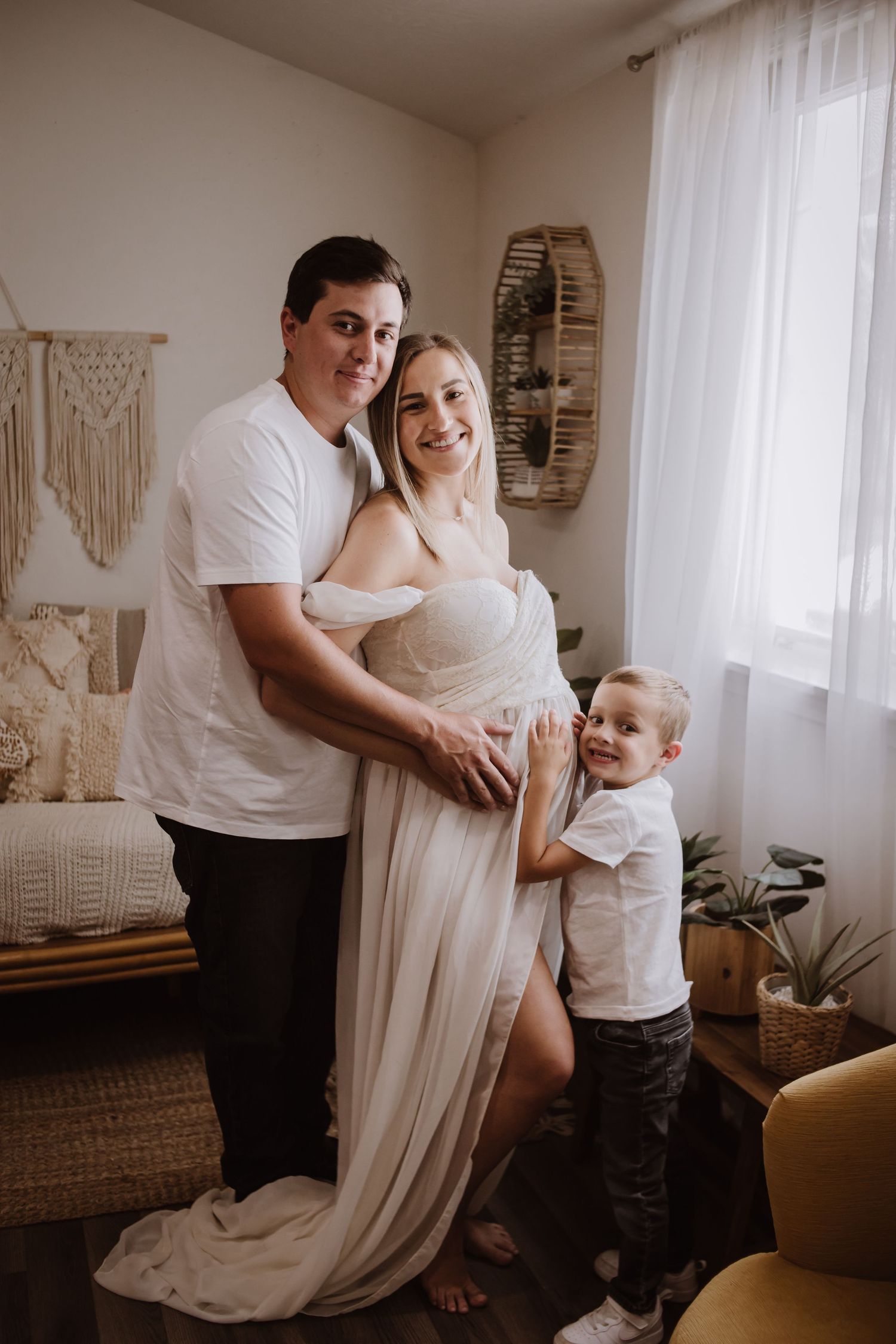 Smiling family maternity photo with pregnant woman in white gown, husband, and young son in a boho-styled room.