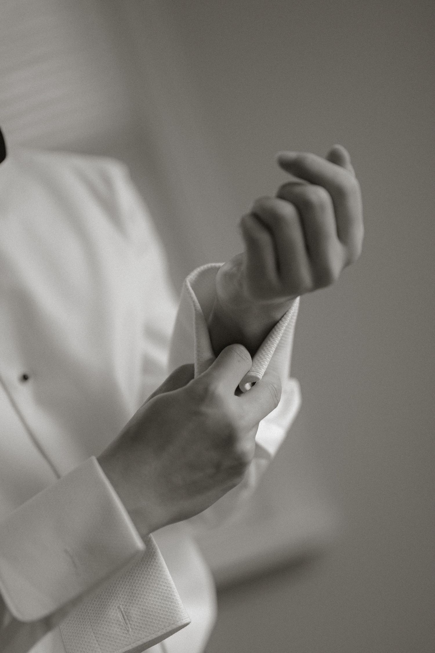 Black and white close-up of groom fastening cufflinks on white tuxedo shirt sleeve.