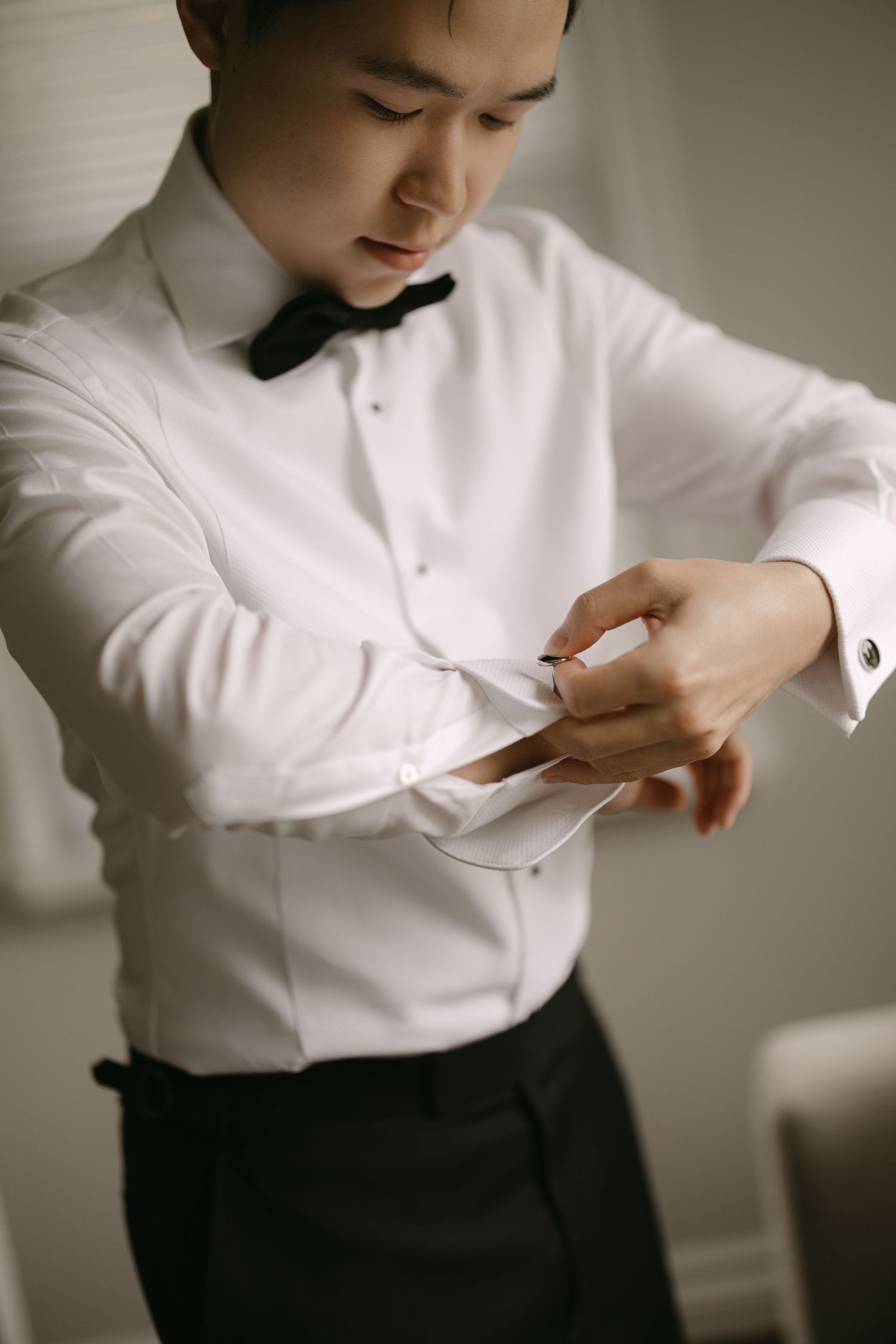 Asian groom in white tuxedo shirt and black bow tie fastening cufflinks while getting ready.