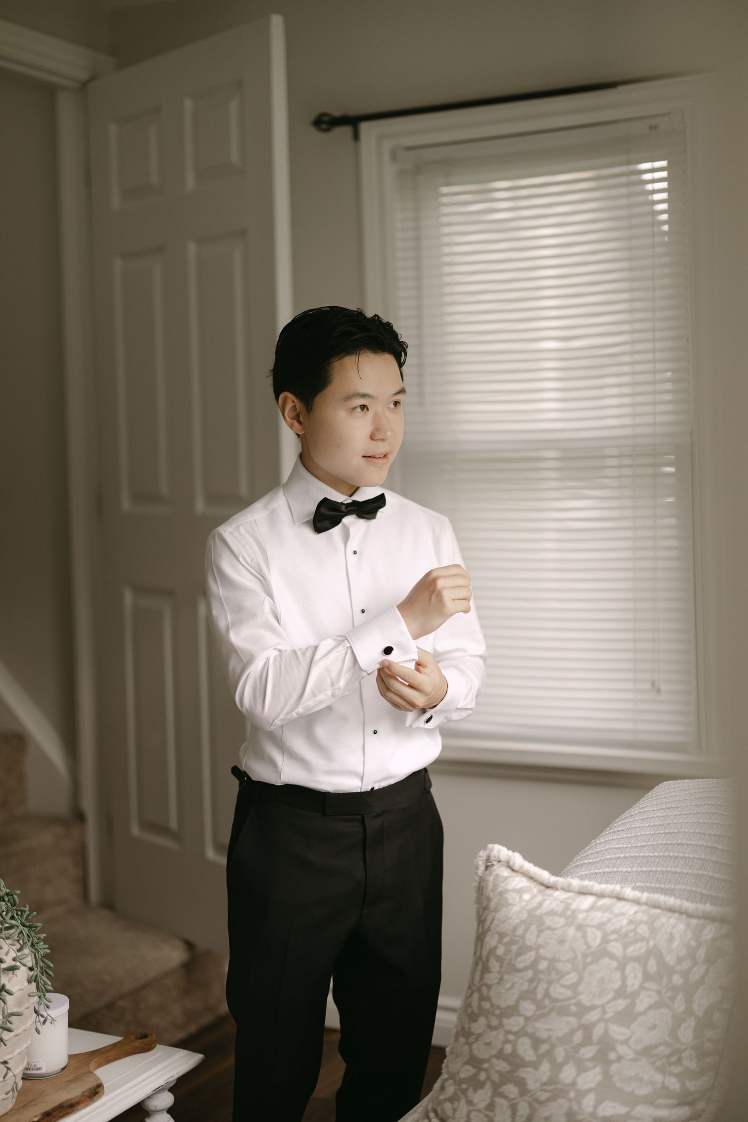 Elegant groom in white shirt and black bow tie adjusting cufflinks by a bright window.