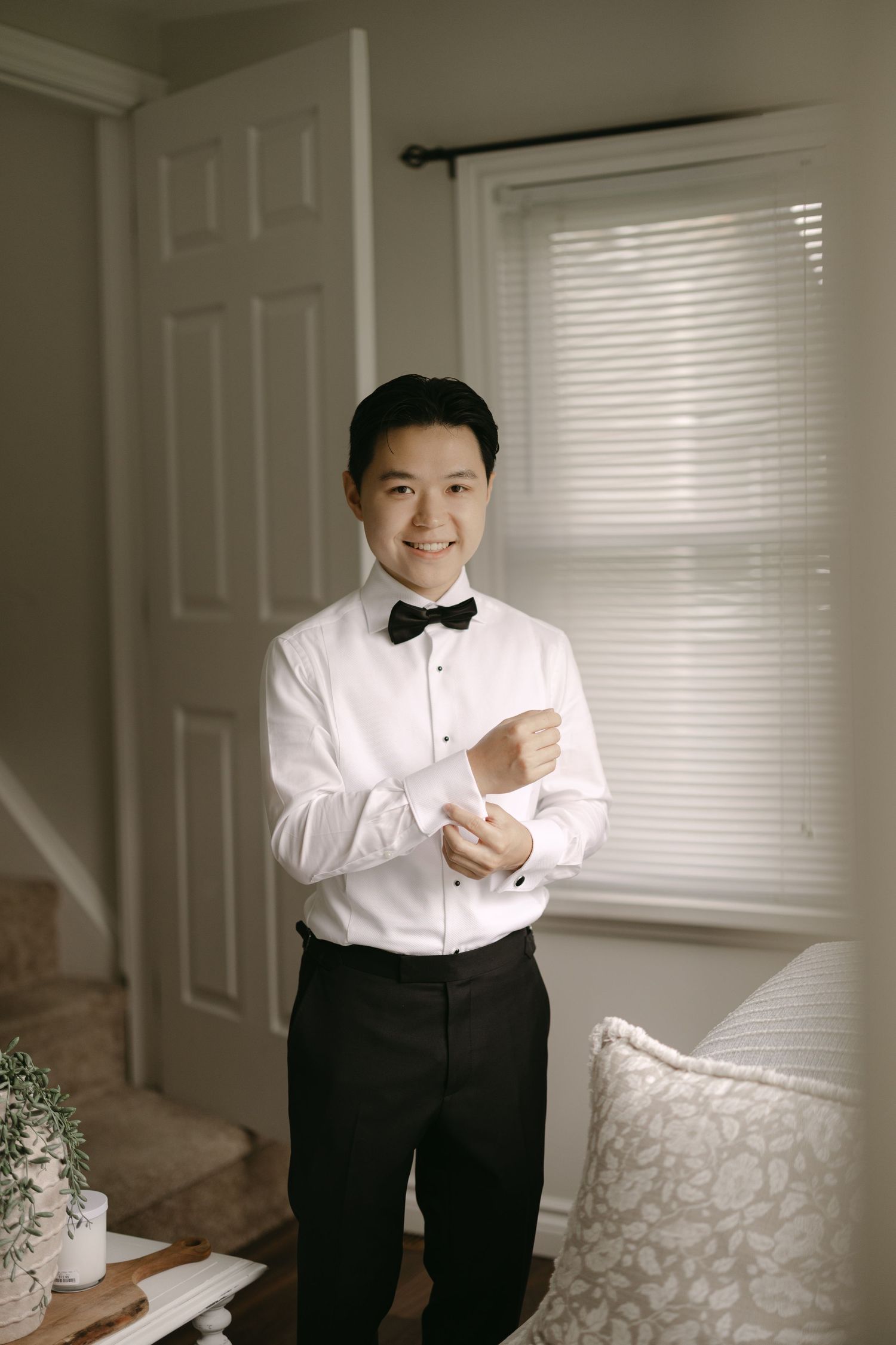 Smiling Asian groom in white tuxedo shirt and black bow tie fastening cufflinks indoors.