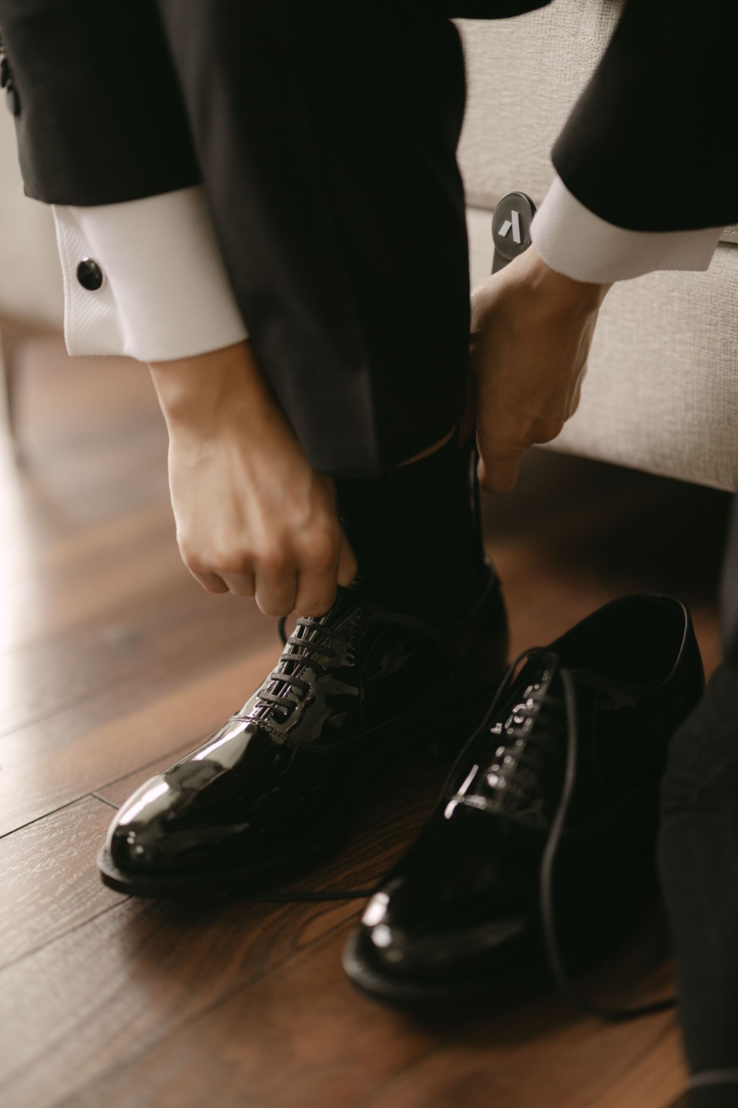 Close-up of groom tying black patent leather dress shoes with white shirt cufflinks visible.
