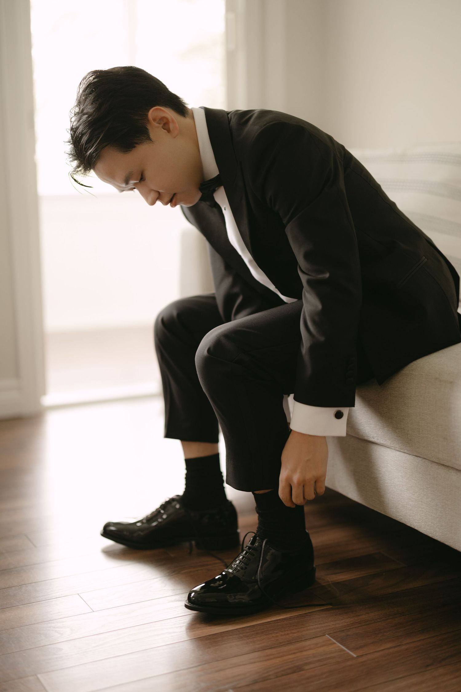 Groom in black tuxedo sitting on sofa putting on black patent leather dress shoes.