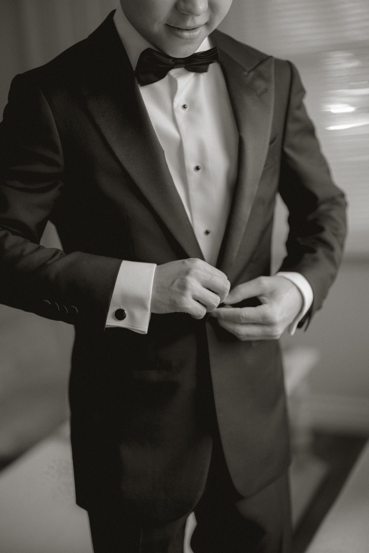 Black and white photo of groom in tuxedo with bow tie buttoning his jacket before wedding ceremony.