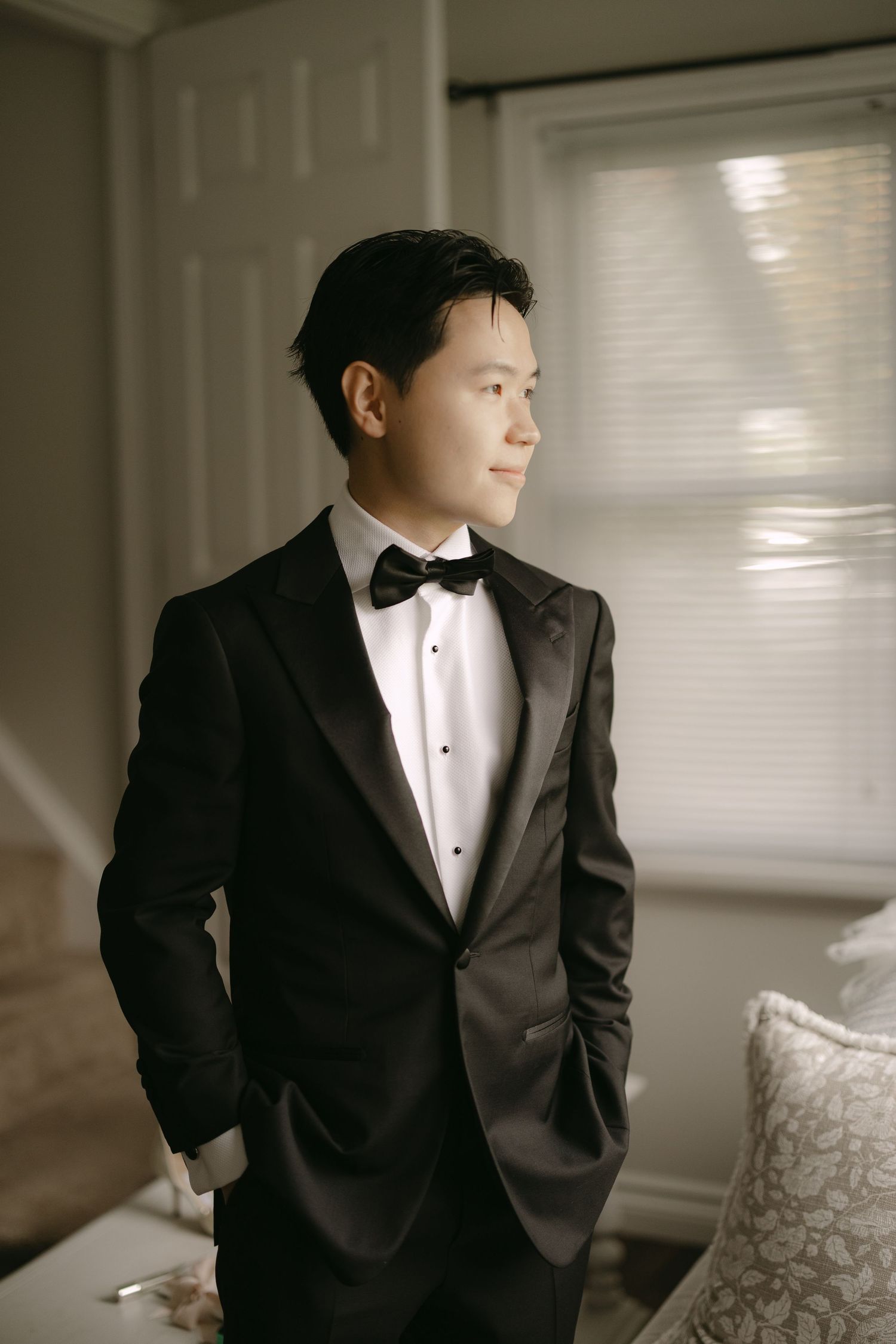 Handsome Asian groom in black tuxedo with bow tie standing by window looking away thoughtfully.
