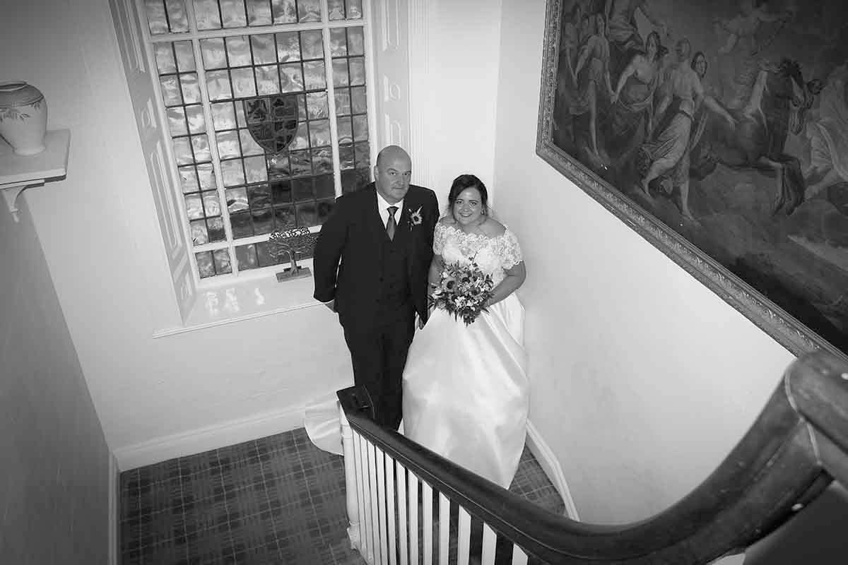 Black and white wedding photo of bride in white gown and groom descending elegant staircase in historic at Blaithwaite House