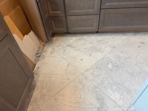 White marble tiles with gray veining installed diagonally on kitchen floor beside dark gray lower cabinets.