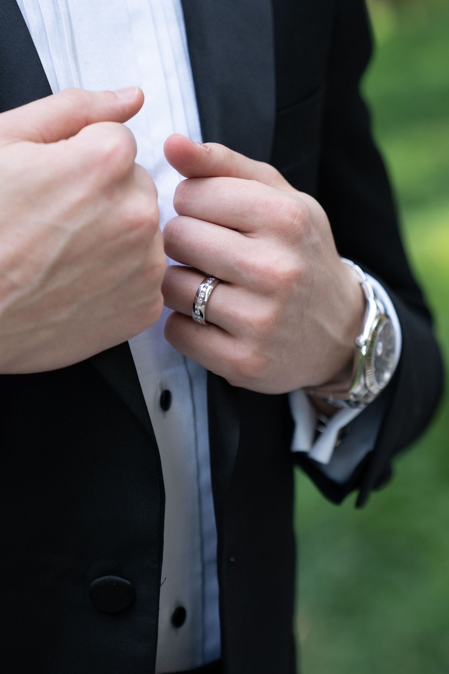 Close-up of groom adjusting tuxedo shirt while wearing wedding band and silver watch.