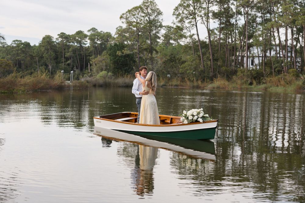 Bride and groom sharing a romantic kiss on a green wooden rowboat on a calm lake surrounded by tall trees.