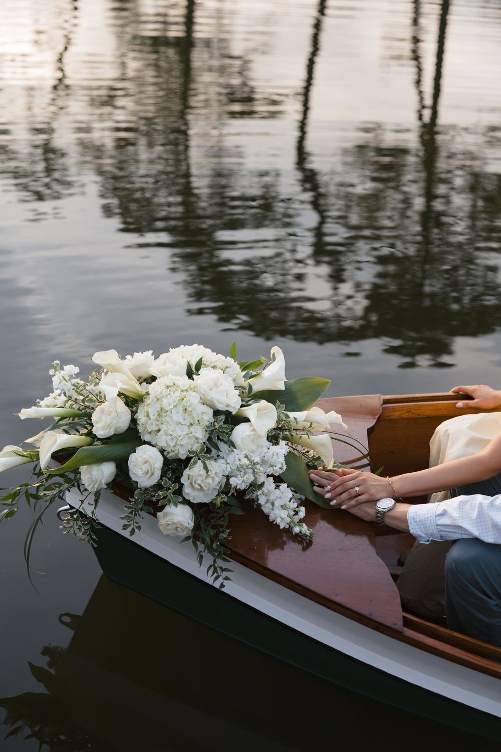Couple holding hands in a wooden boat with a large white floral bouquet featuring roses and calla lilies on calm water.