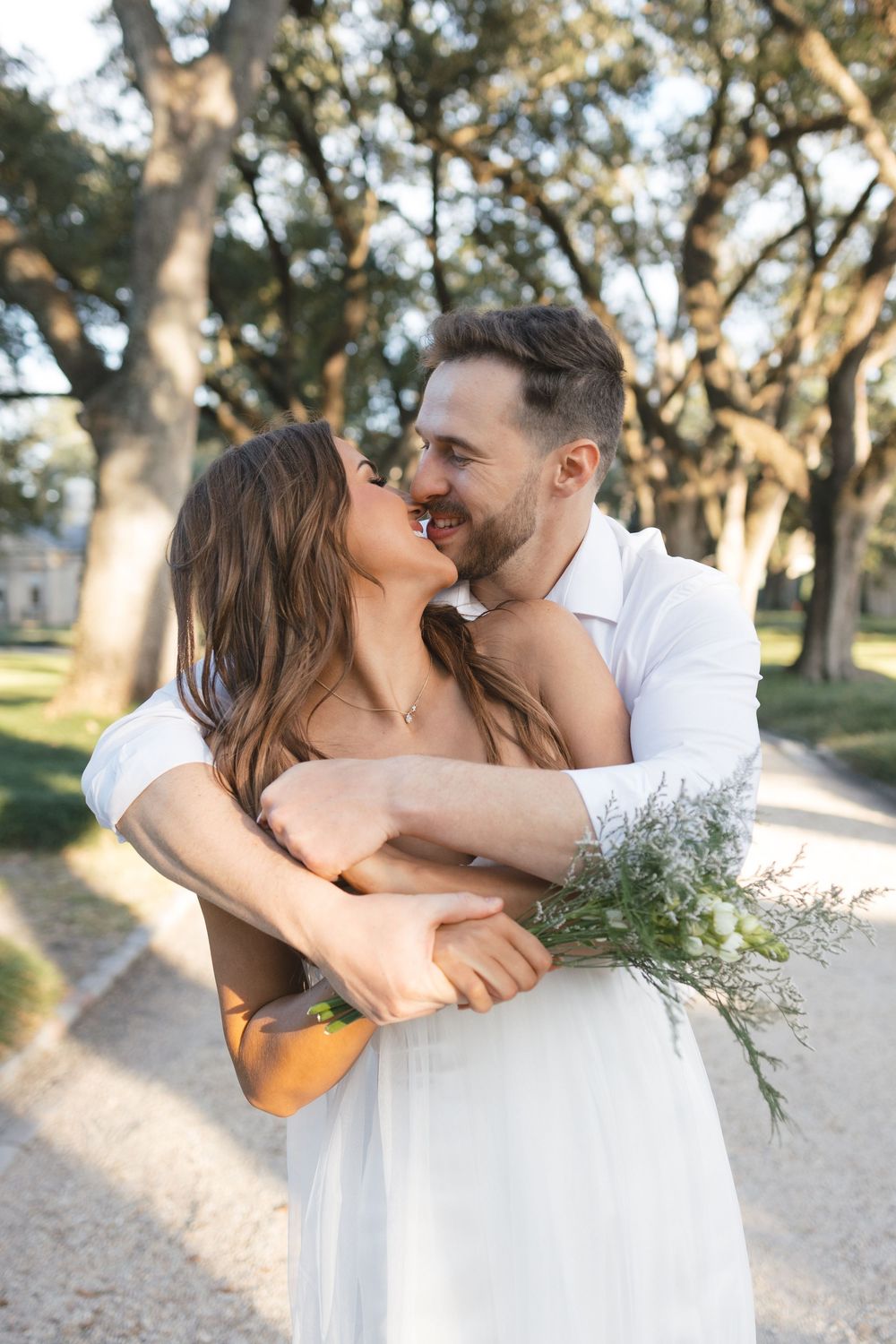 Happy couple embracing and almost kissing outdoors among tall trees, woman holding wildflower bouquet.