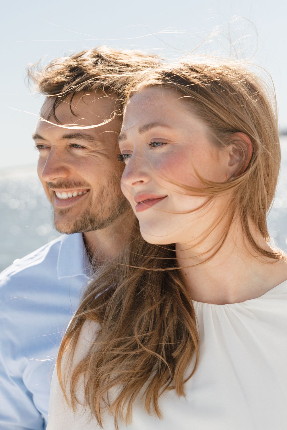 A smiling couple with windswept hair poses outdoors on a sunny day, the woman leaning close to the man in a tender moment.