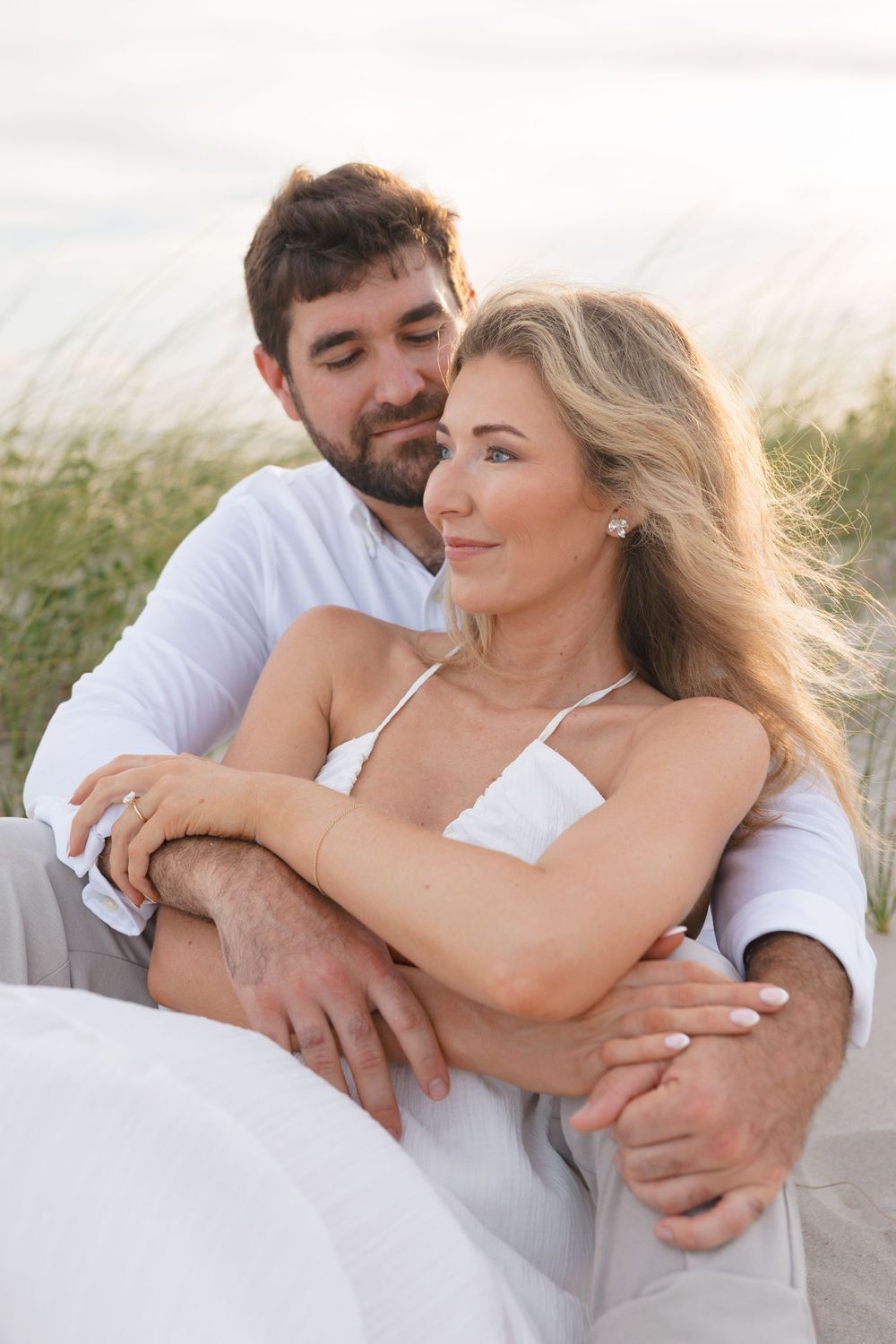 Couple sitting together on beach dunes at sunset, man embracing woman from behind in white outfits.