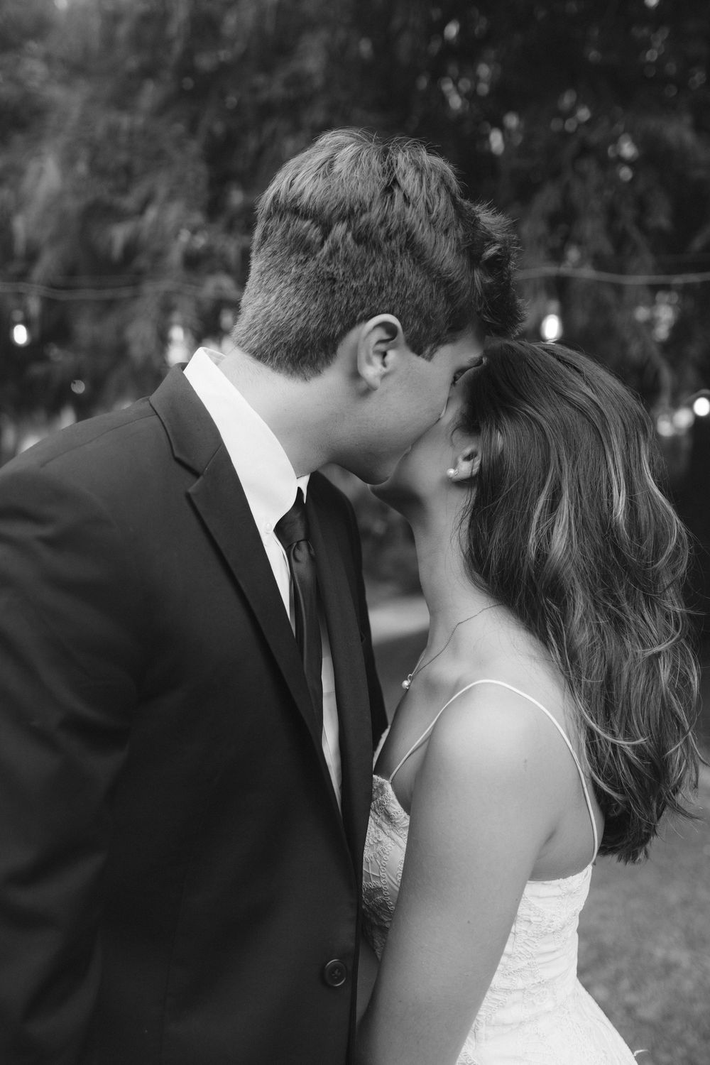 Black and white photo of bride and groom sharing a romantic kiss outdoors under soft string lights.