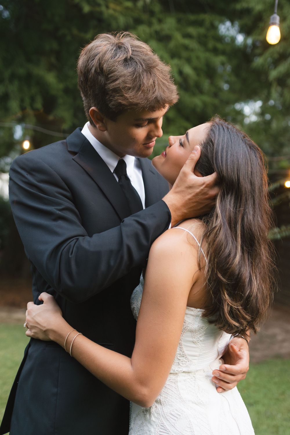 Groom tenderly holding brides face before a kiss under glowing string lights in an outdoor garden setting.